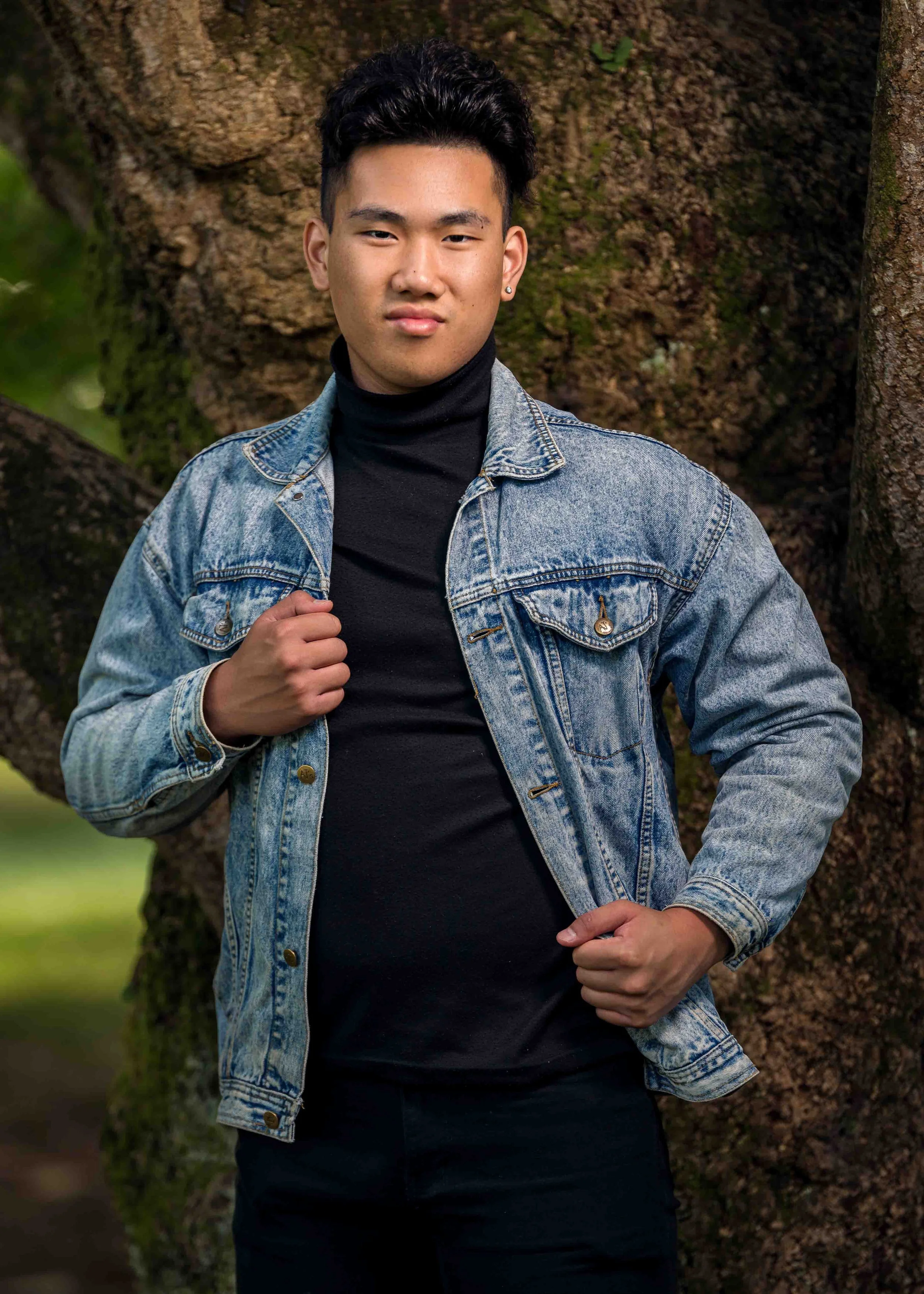Young man wearing a denim jacket and black turtleneck standing outdoors in front of a tree.