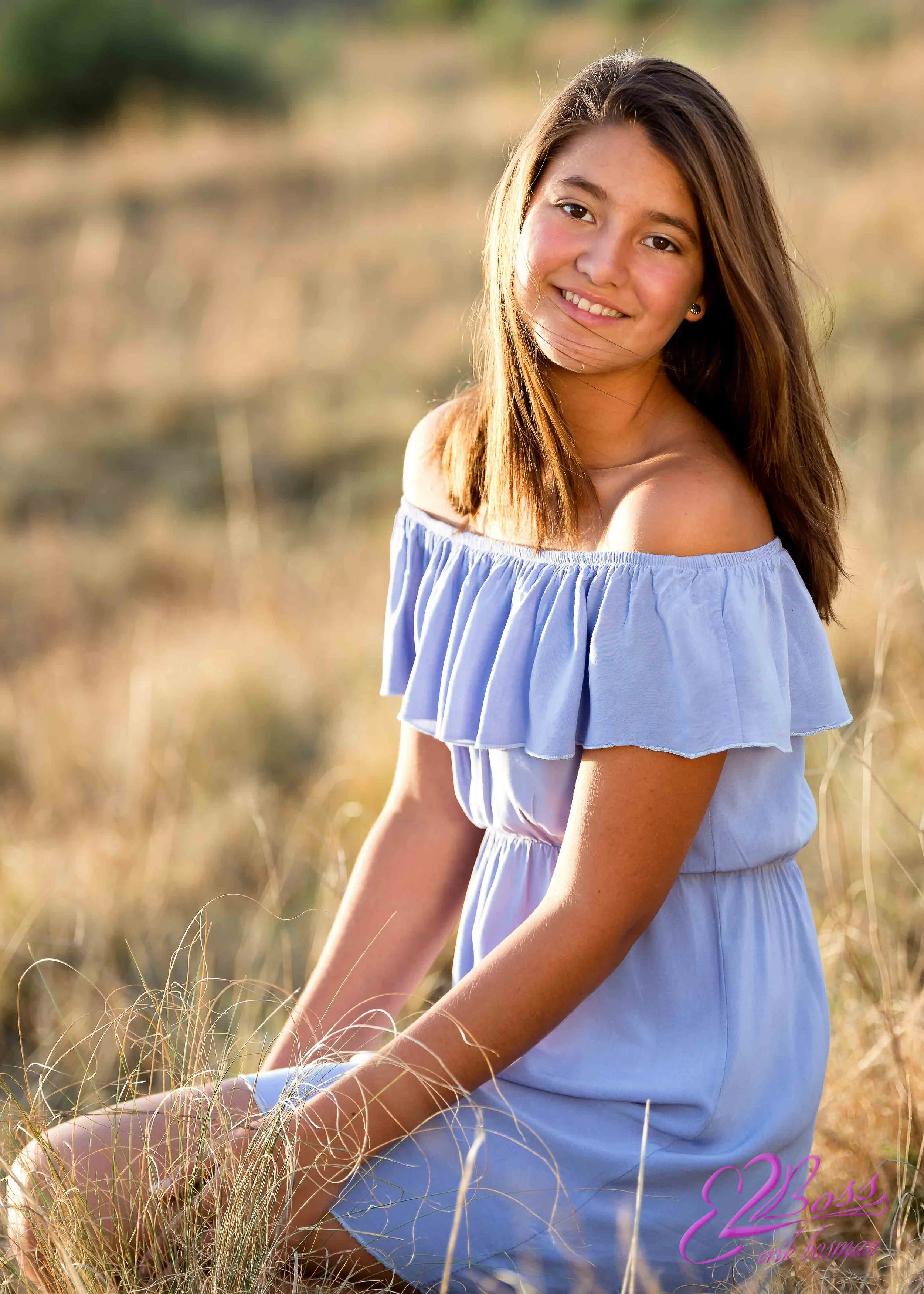A young woman with long brown hair sitting in a field of dry grass, smiling at the camera, wearing a light blue off-shoulder dress during golden hour.