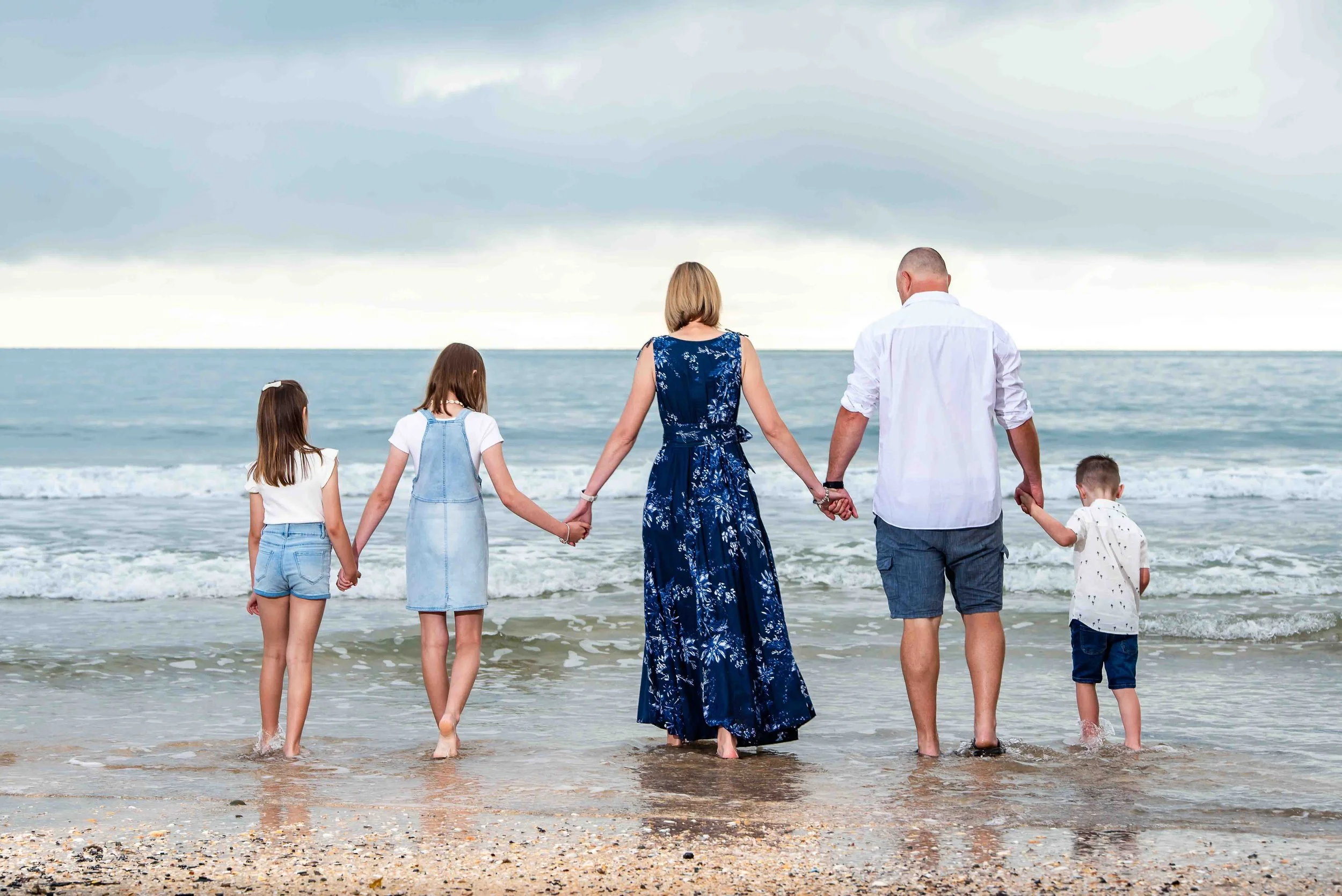 A family of five holding hands walking into the ocean on a cloudy day.