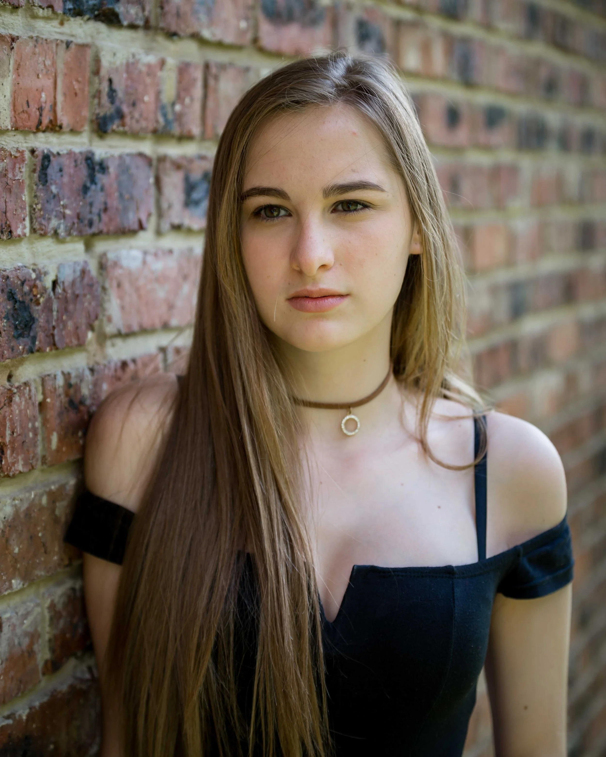 A young woman with long brown hair leaning against a brick wall, looking to the side with a neutral expression, wearing a black off-shoulder top and a choker necklace.