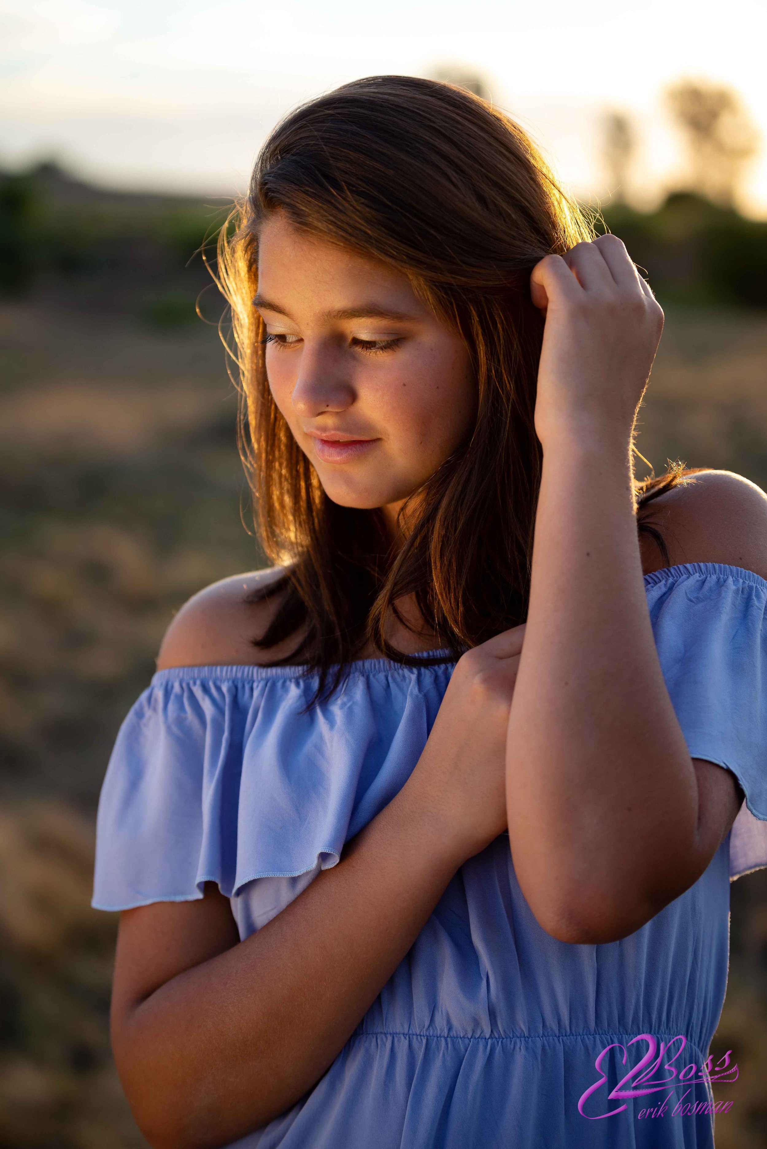 A young woman with brown hair and fair skin stands outdoors during sunset, wearing a light blue off-shoulder top, with her eyes closed and her hand touching her hair.