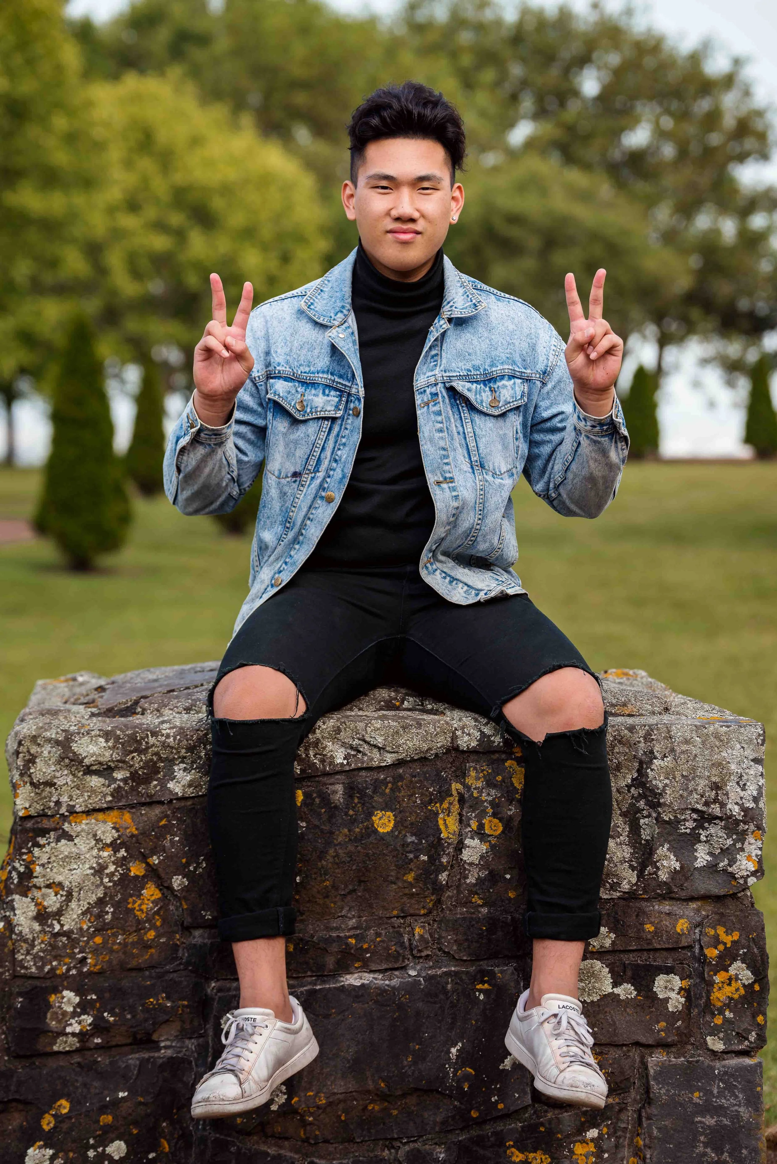 Young man sitting on a stone wall outdoors, making peace signs with both hands, wearing a denim jacket, black ripped jeans, and white sneakers, with trees in the background.