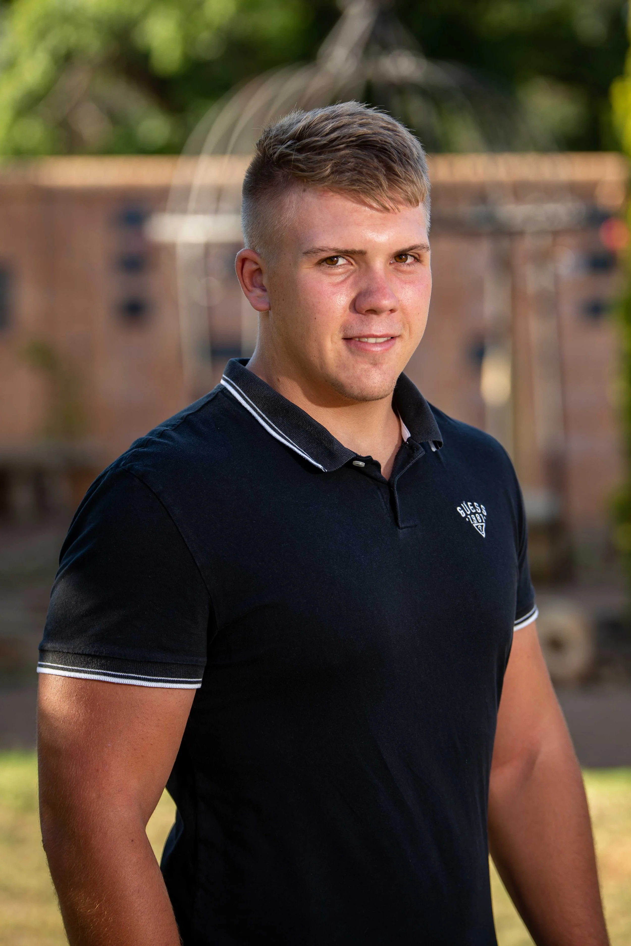 A young man with short, styled hair and wearing a black Guess polo shirt, standing outdoors in a backyard with greenery and a brick fire pit in the background.