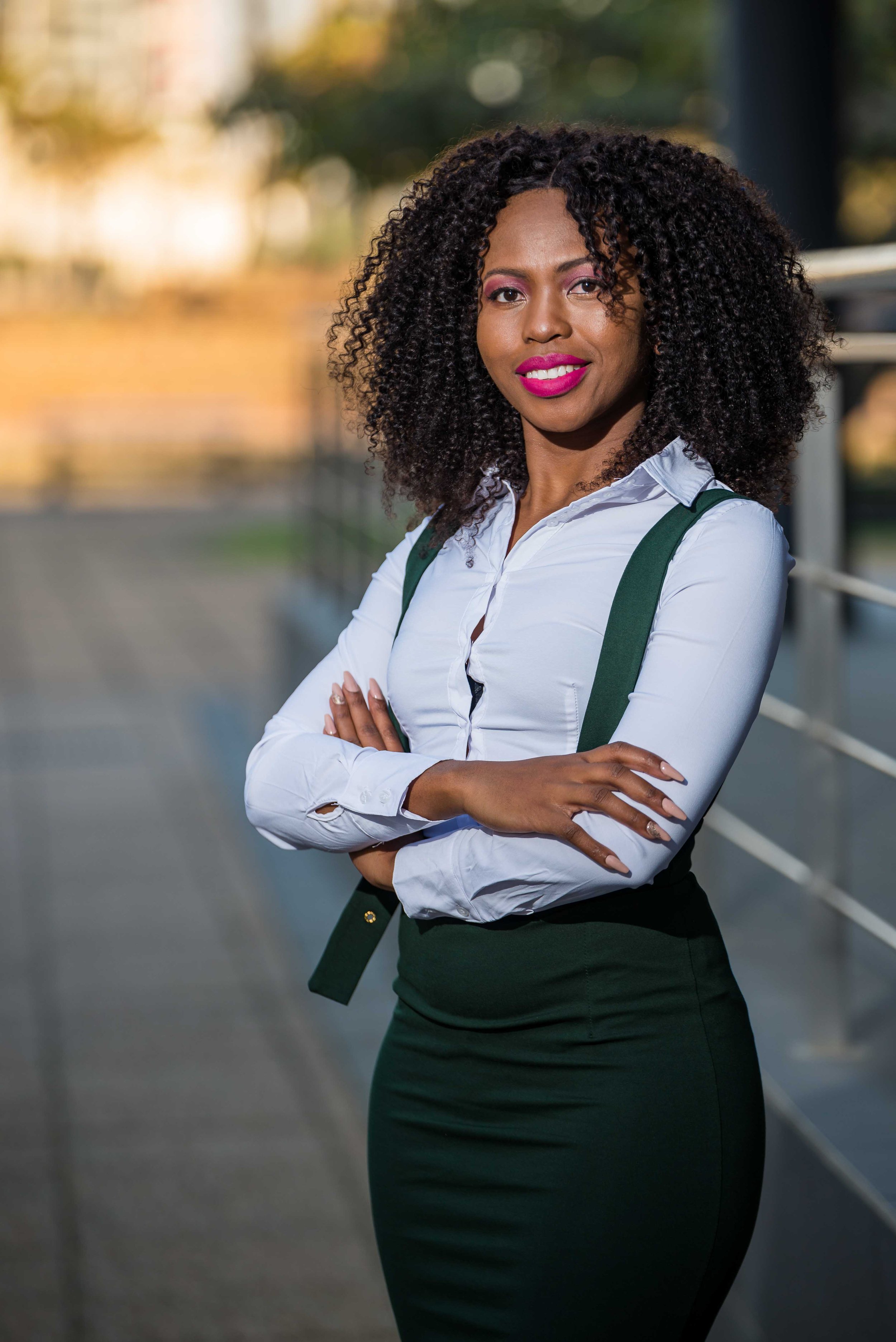 A confident woman standing outdoors with arms crossed, wearing a white shirt and dark green skirt, with a backpack strap visible on her shoulder.