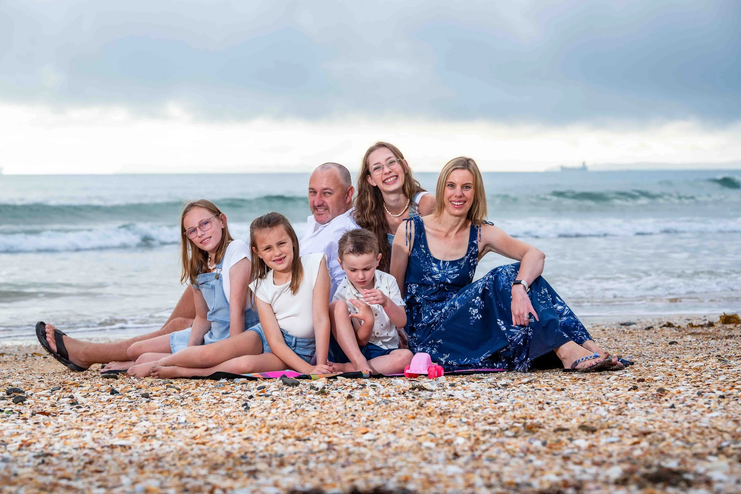 A family of seven sitting on a pebble beach with the ocean in the background, smiling and enjoying their time together.