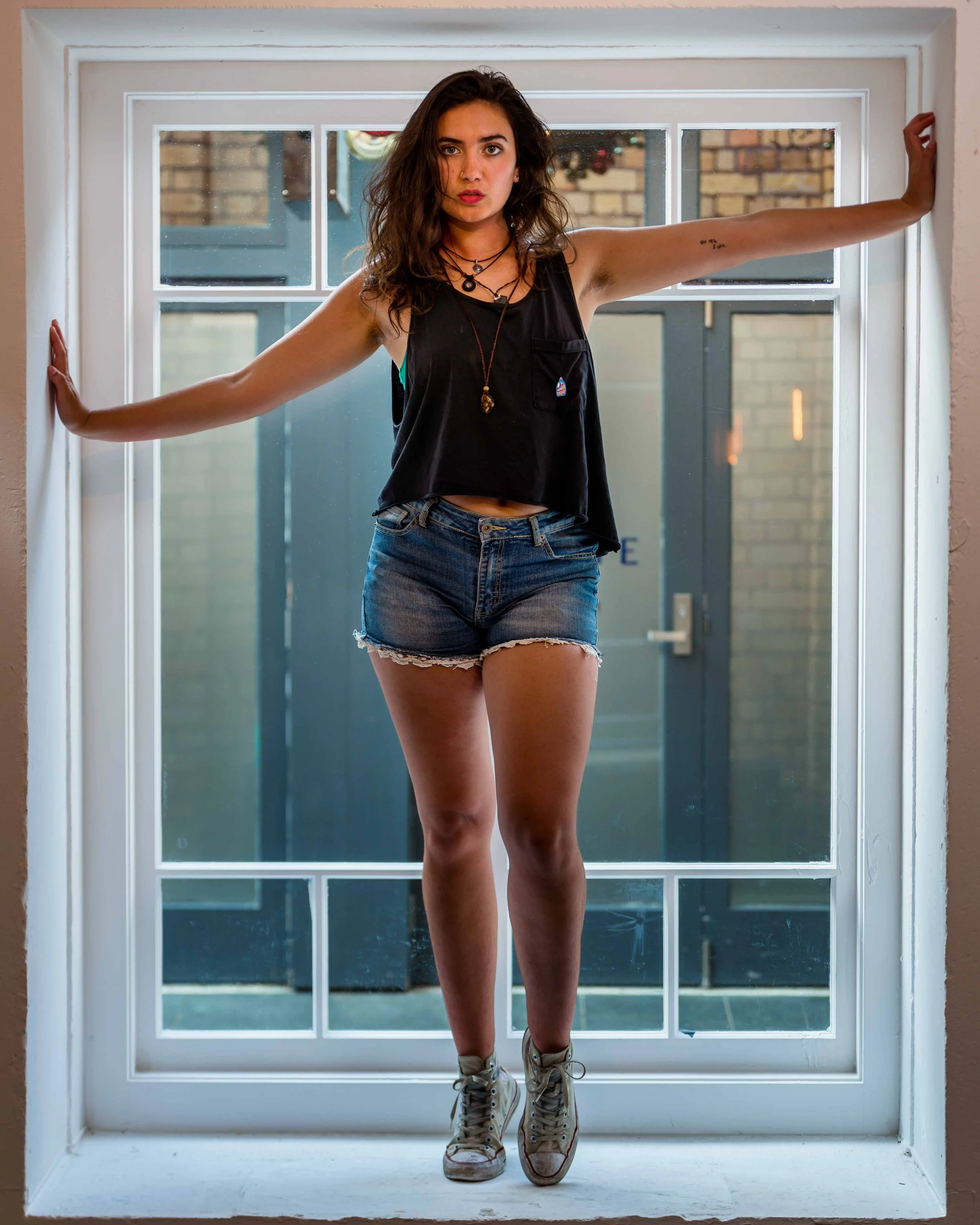 A young woman with wavy brown hair standing in front of a window, wearing a black sleeveless top, denim shorts, and sneakers, with her arms stretched out touching the window frame.