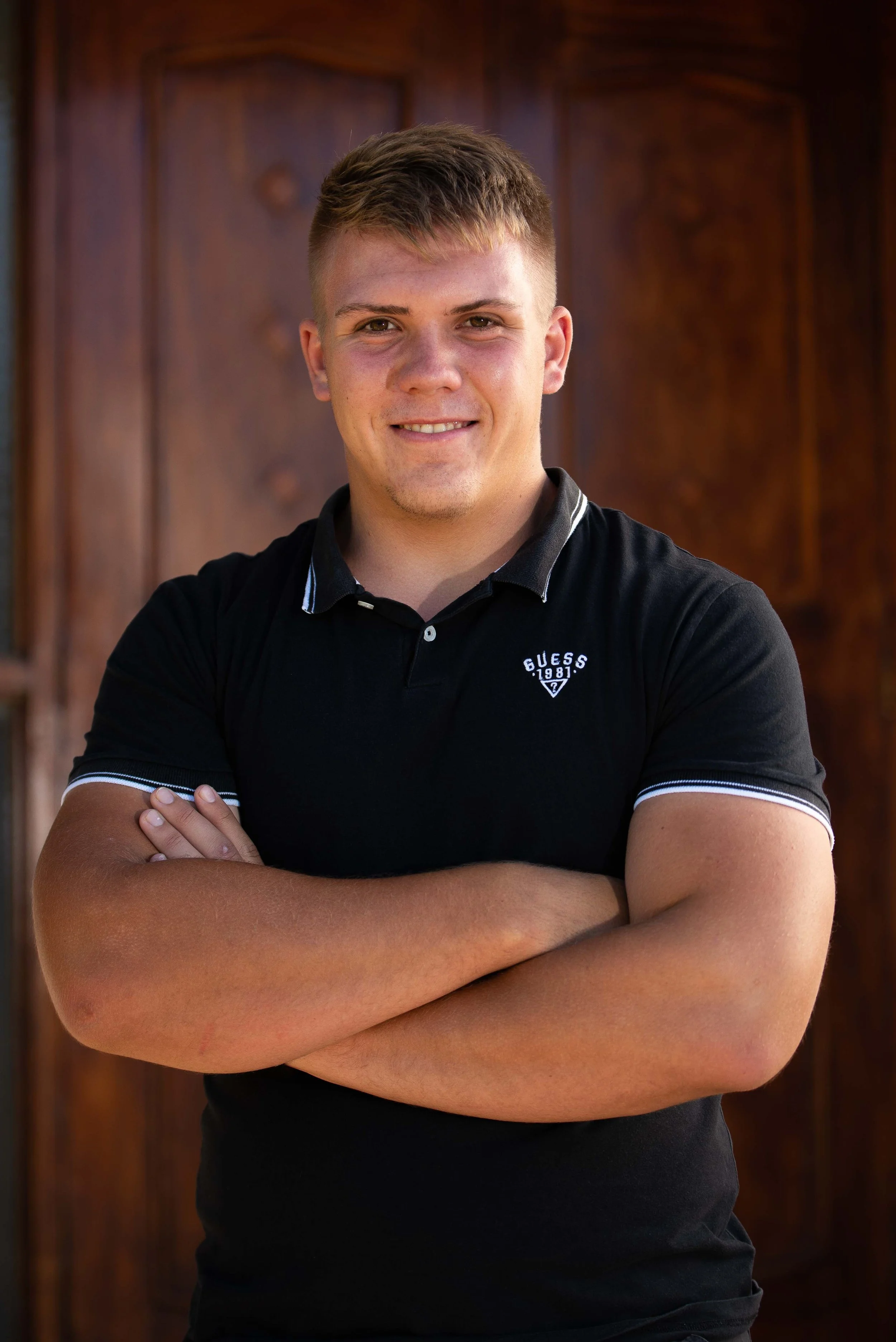 A young man with light skin and short brown hair, smiling with arms crossed, wearing a black Guess polo shirt, standing in front of a wooden background.