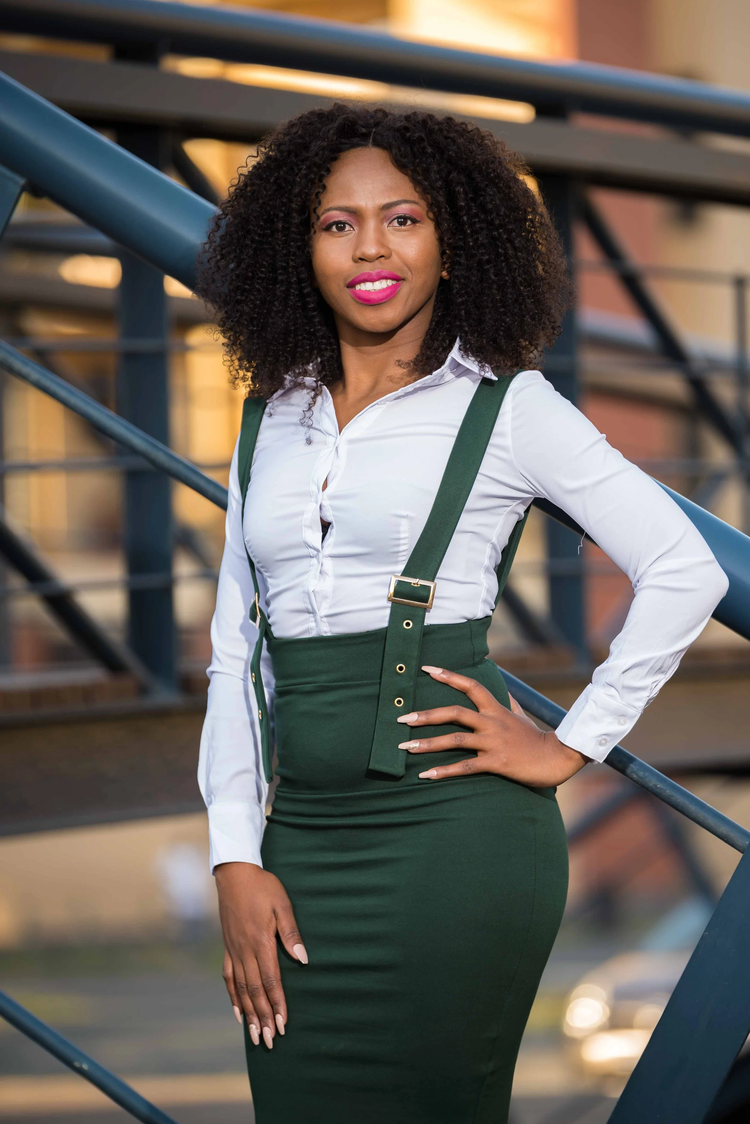 A woman with curly hair wearing a white button-up shirt and a green skirt with suspenders, standing outdoors near a staircase and smiling at the camera.
