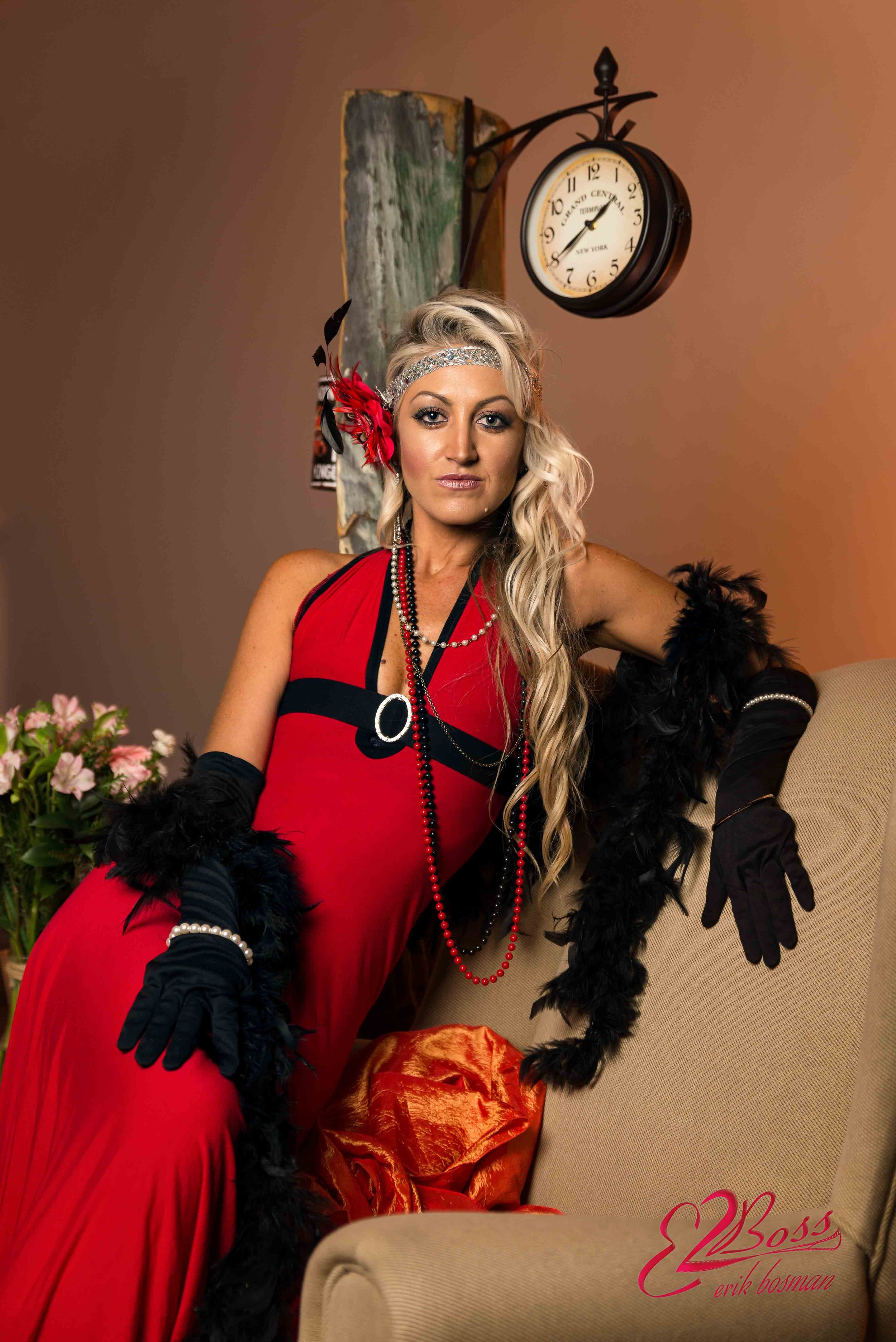 A woman with long blonde hair wearing a red dress, black gloves, and black feathered accessories, seated on a beige chair in an interior setting with a clock and artwork on the wall behind her.