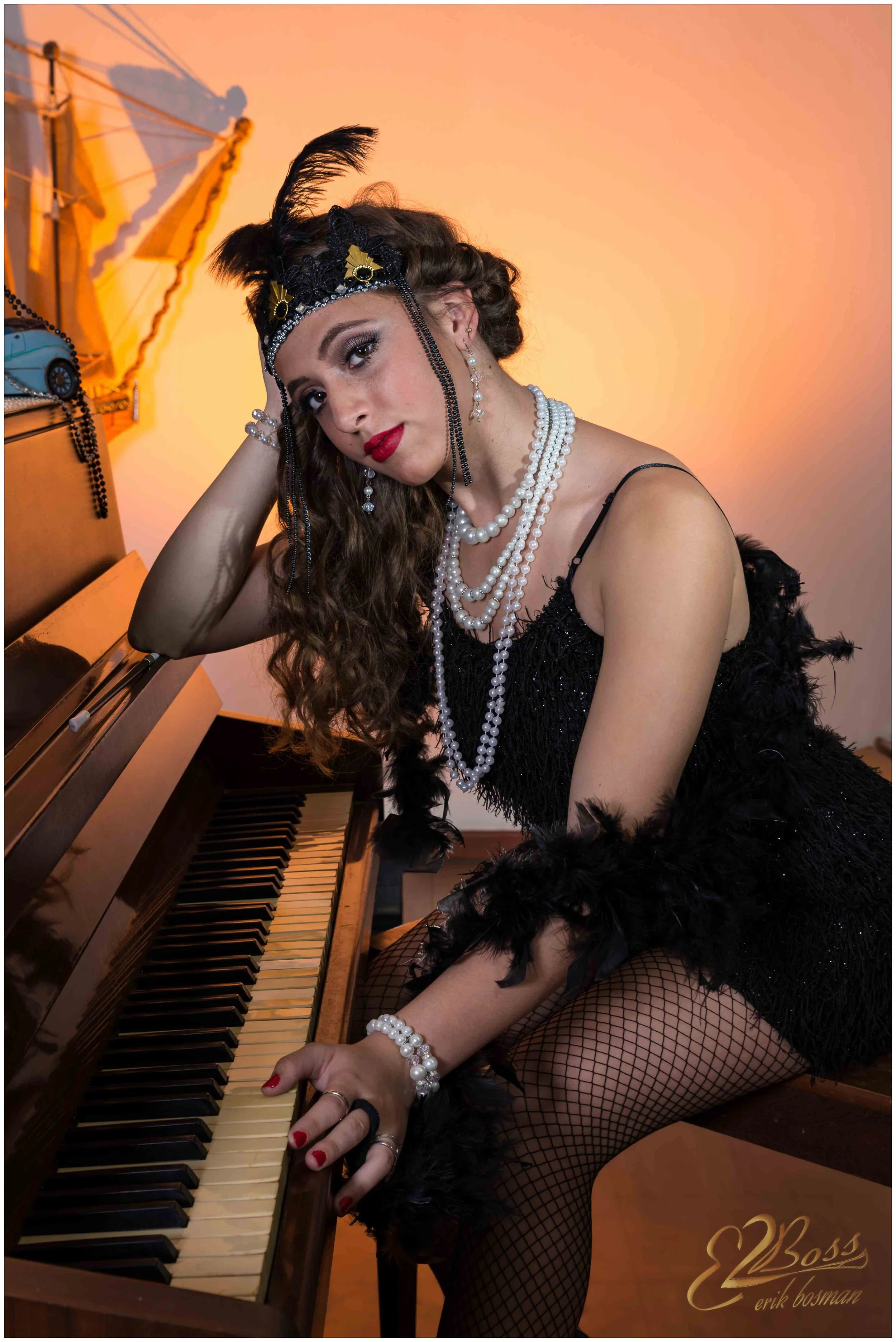 A woman dressed in a black flapper-style dress with feather accessories, wearing pearls and fishnet stockings, sitting at a vintage piano with an orange background and a small model ship on a table behind her.