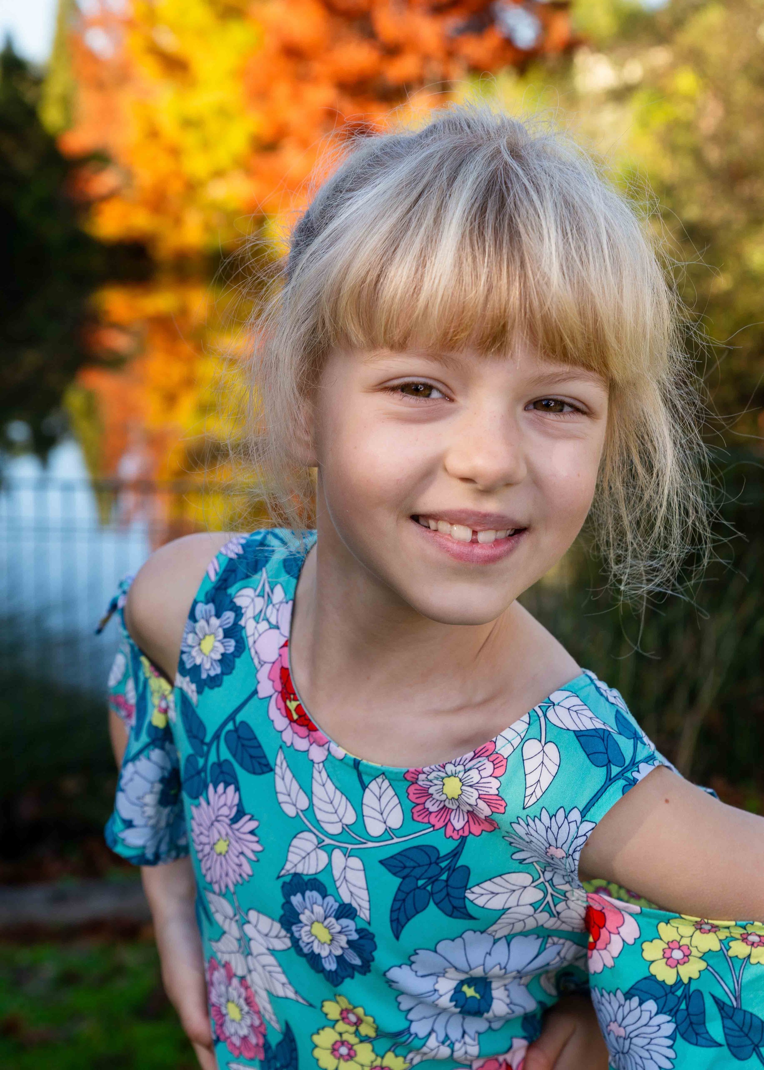 A young girl with blonde hair and a floral dress smiling outdoors with autumn foliage in the background.