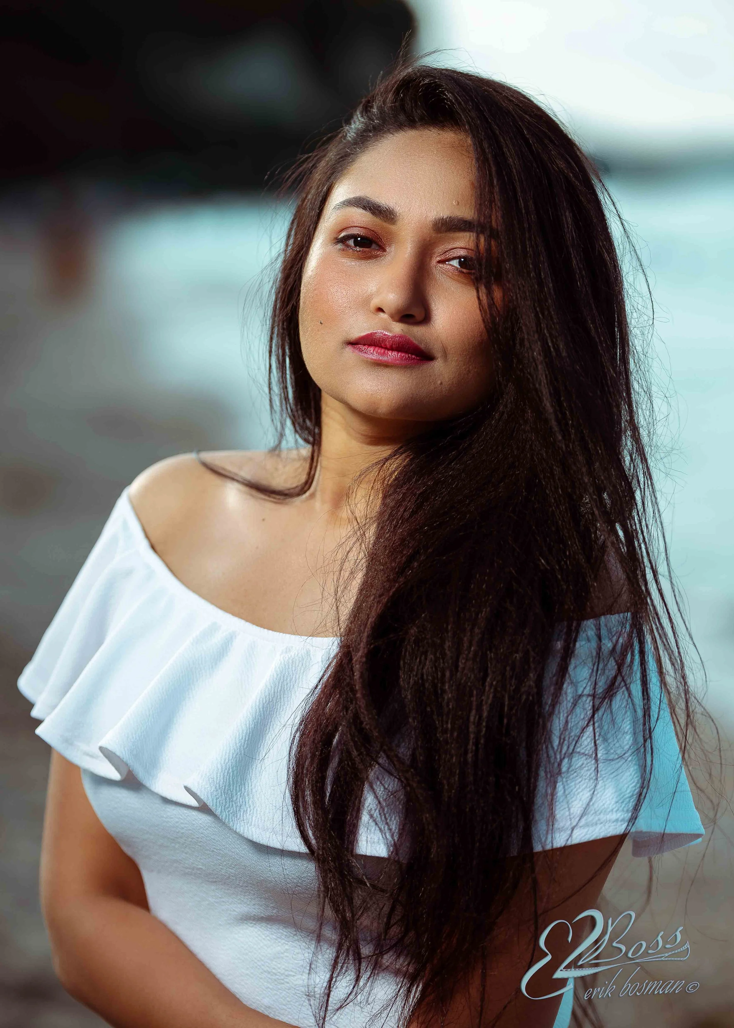 A woman with long dark hair wearing a white off-the-shoulder top, standing outdoors near water, with a confident expression.