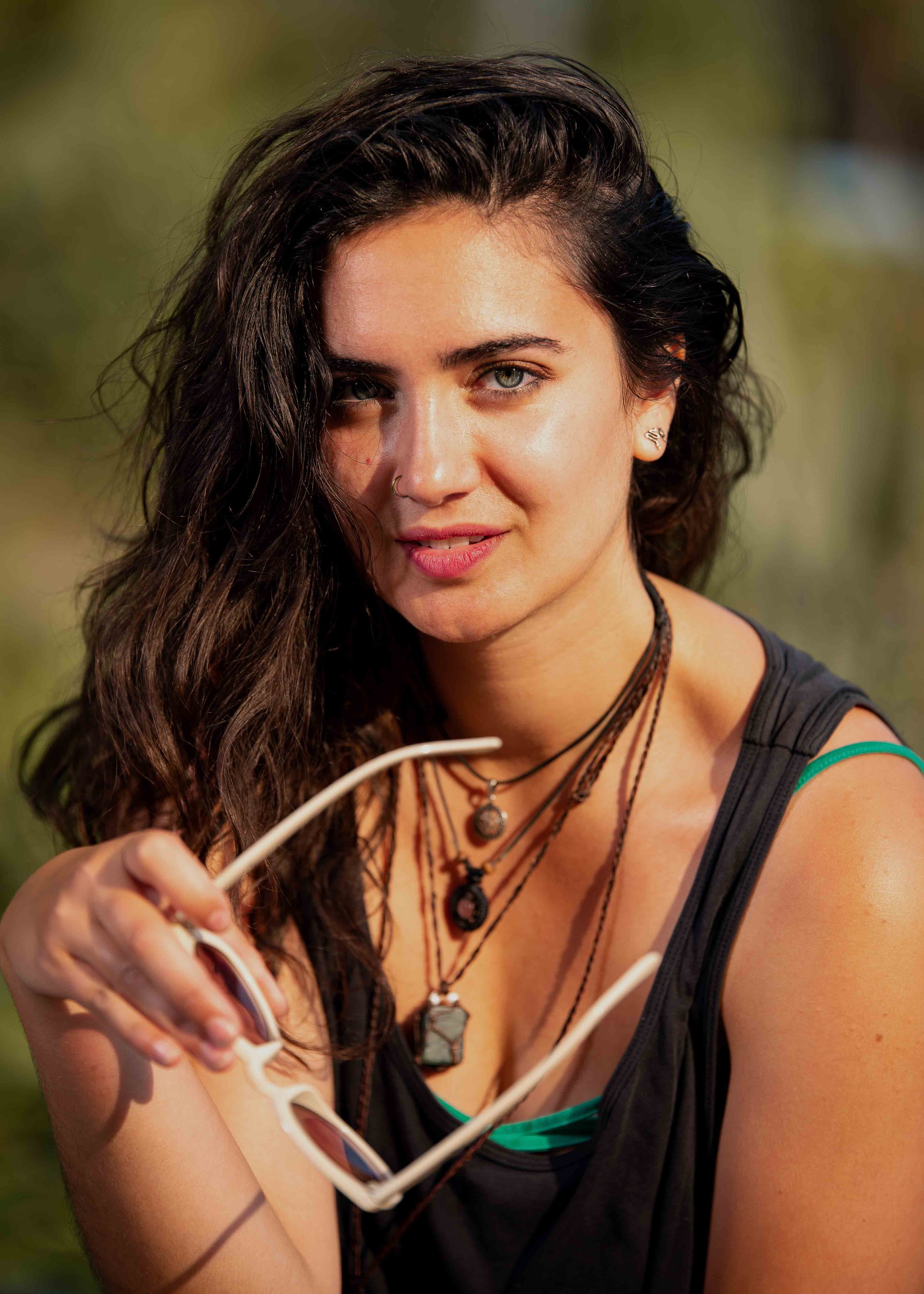 Young woman with dark, wavy hair holding sunglasses, wearing layered necklaces, earrings, and a black tank top with green straps, outdoors.