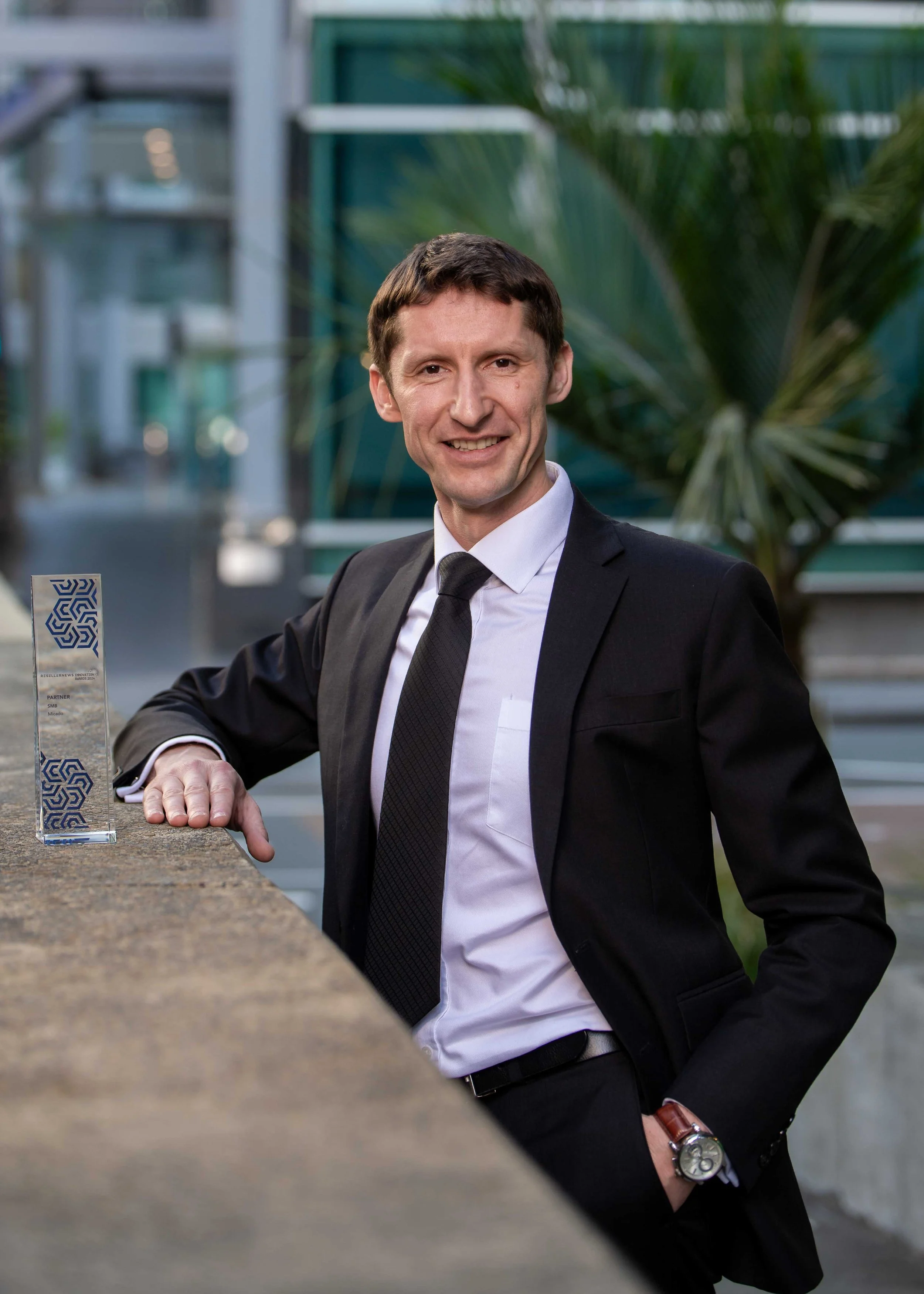 A man in a black suit, white shirt, and black tie posing outdoors next to a stone table with a glass award, smiling at the camera, with a modern building and green plants in the background.