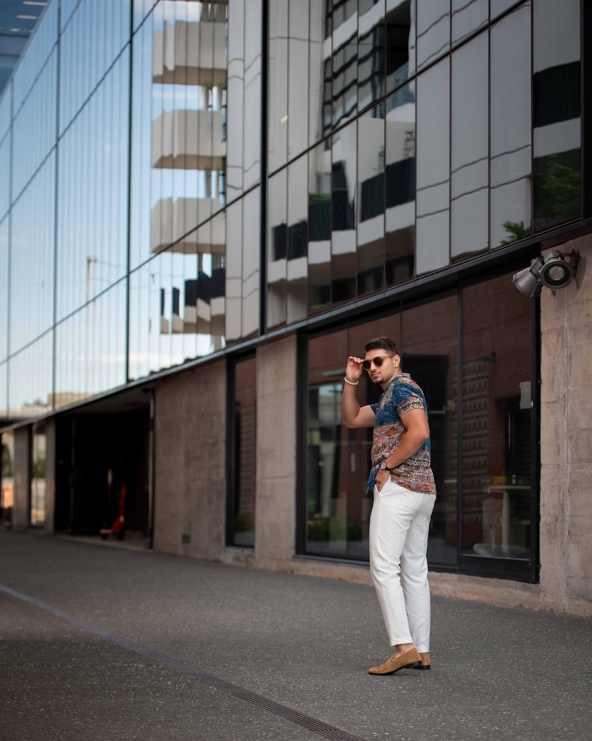 A man in a patterned short-sleeve shirt, white pants, and tan shoes stands outside a modern glass building, adjusting his sunglasses with one hand and placing the other hand in his pocket.