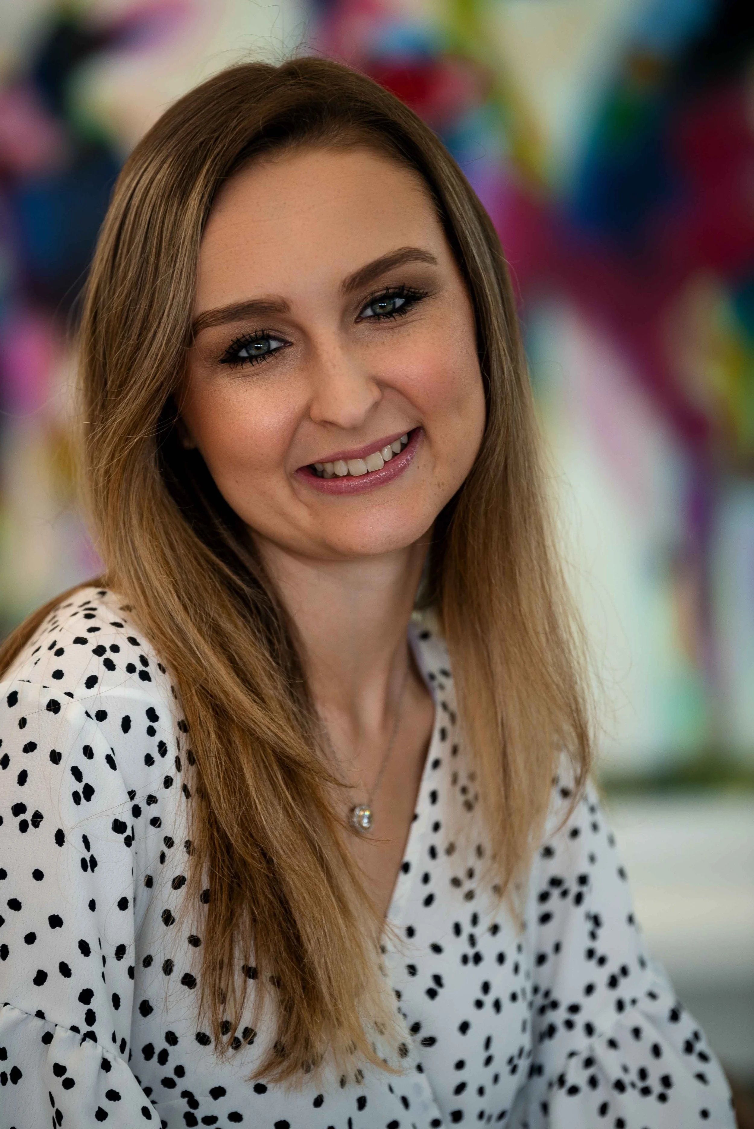 A young woman with long, light brown hair, blue eyes, and a friendly smile, wearing a white blouse with black polka dots and a silver pendant necklace, standing in front of a colorful, blurred background.