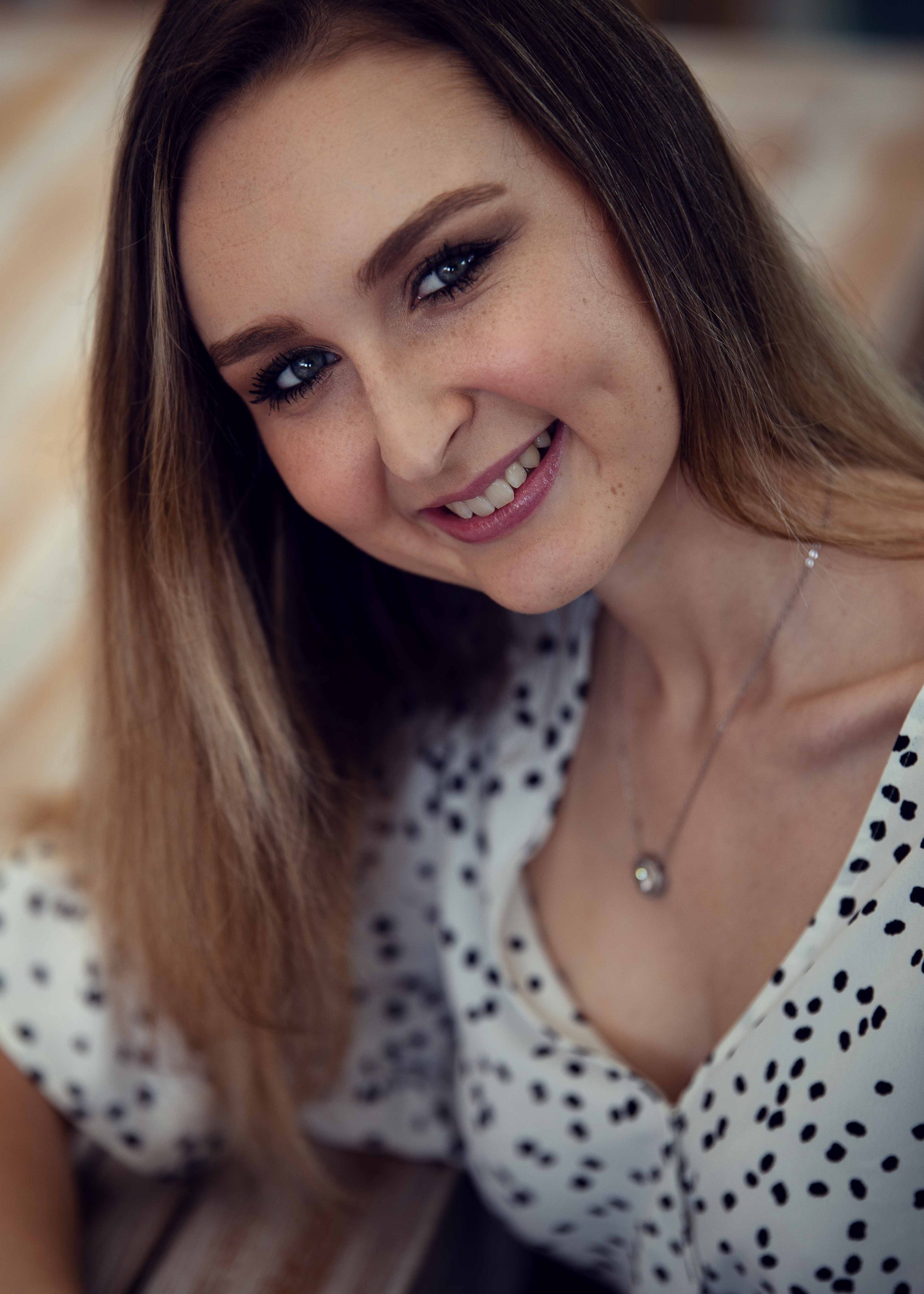 Close-up portrait of a smiling woman with long light brown hair, wearing a white blouse with black polka dots and a silver necklace with a pendant.