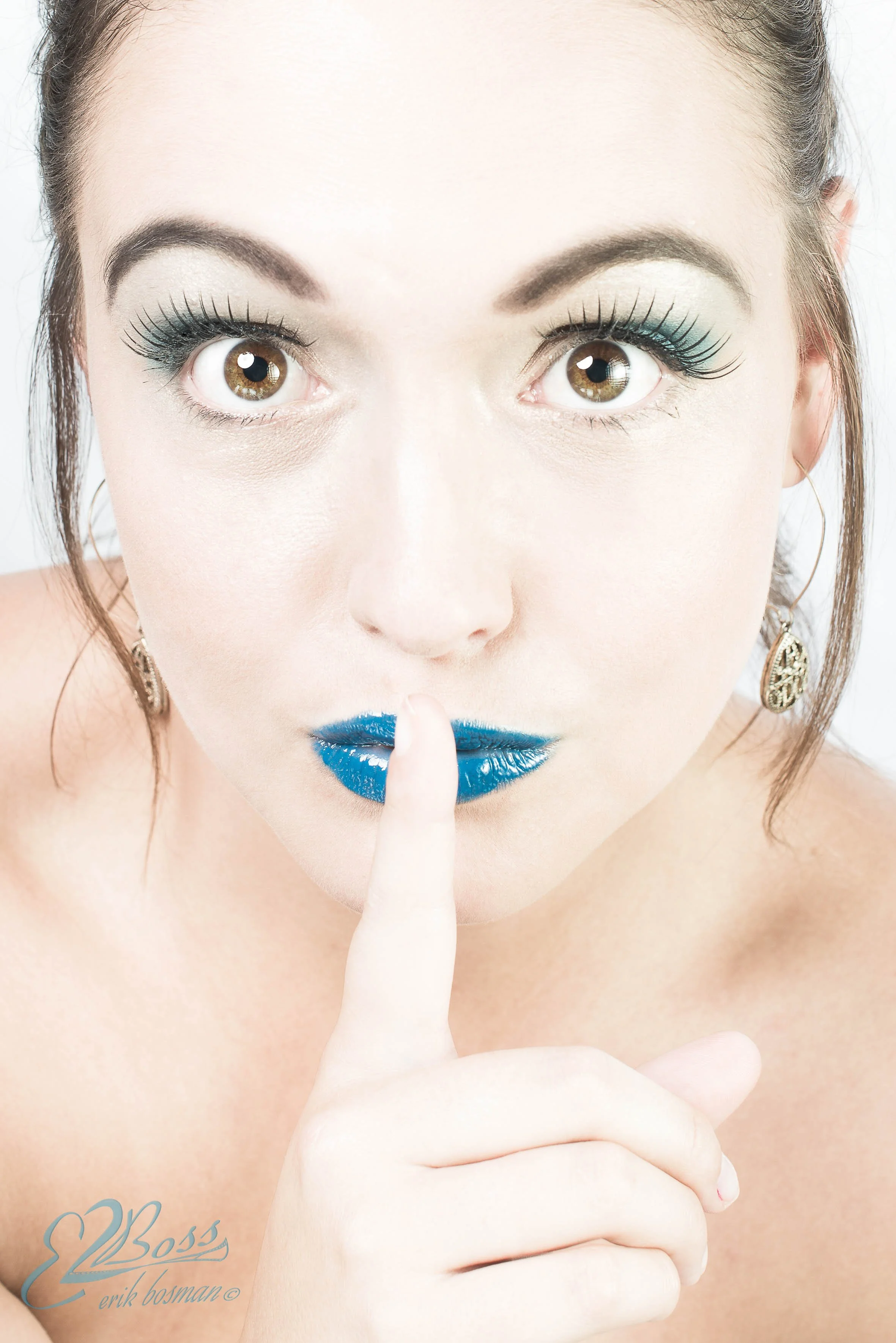 A woman with bright makeup, turquoise lipstick, day-glow eyeshadow, and long eyelashes, holding her finger to her lips in a shushing gesture.