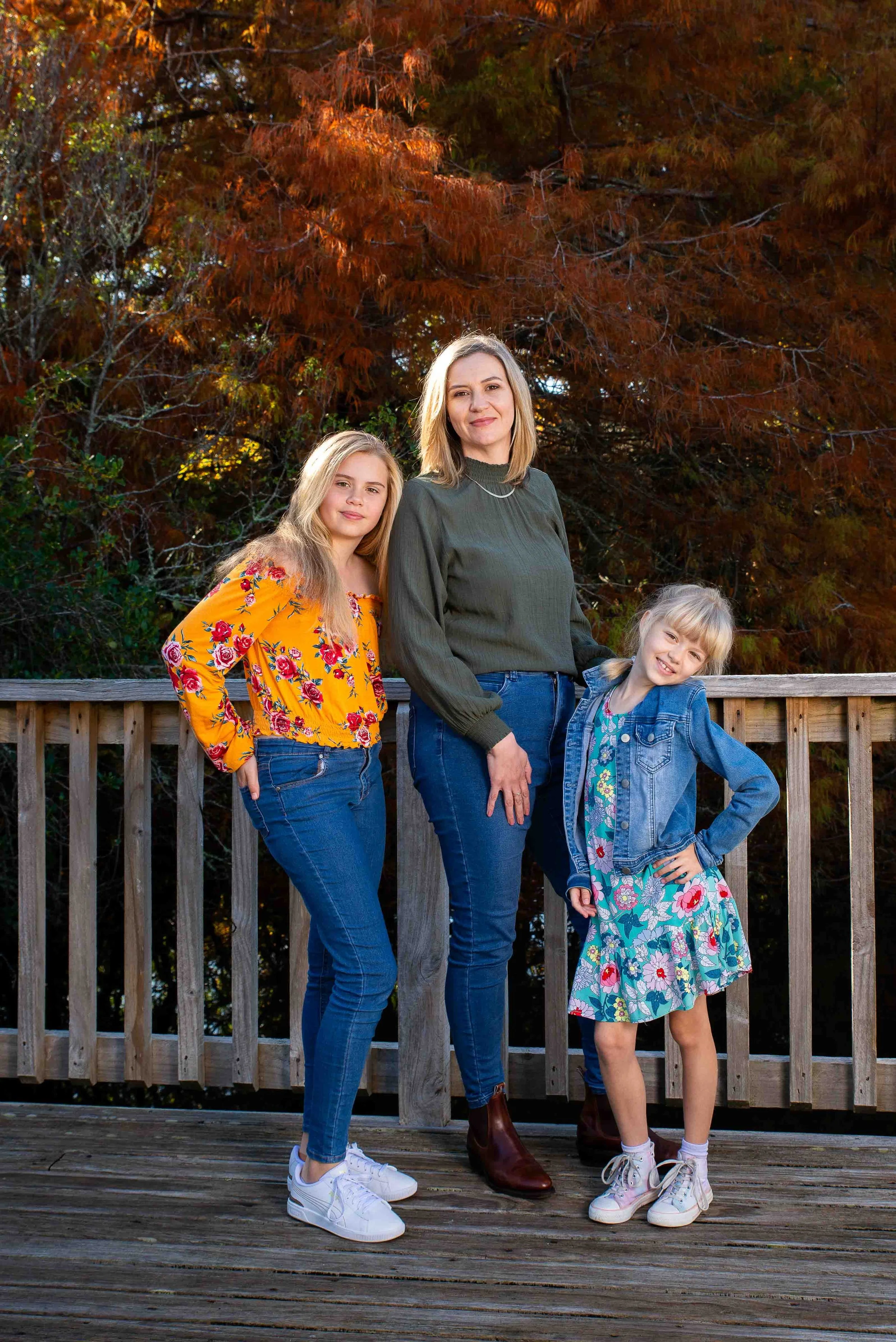 Three females standing on a wooden deck with a fence, outdoors during autumn, with trees with orange leaves in the background. An adult woman is in the middle with her arm around a young girl in a floral dress and denim jacket on the right, and anoth