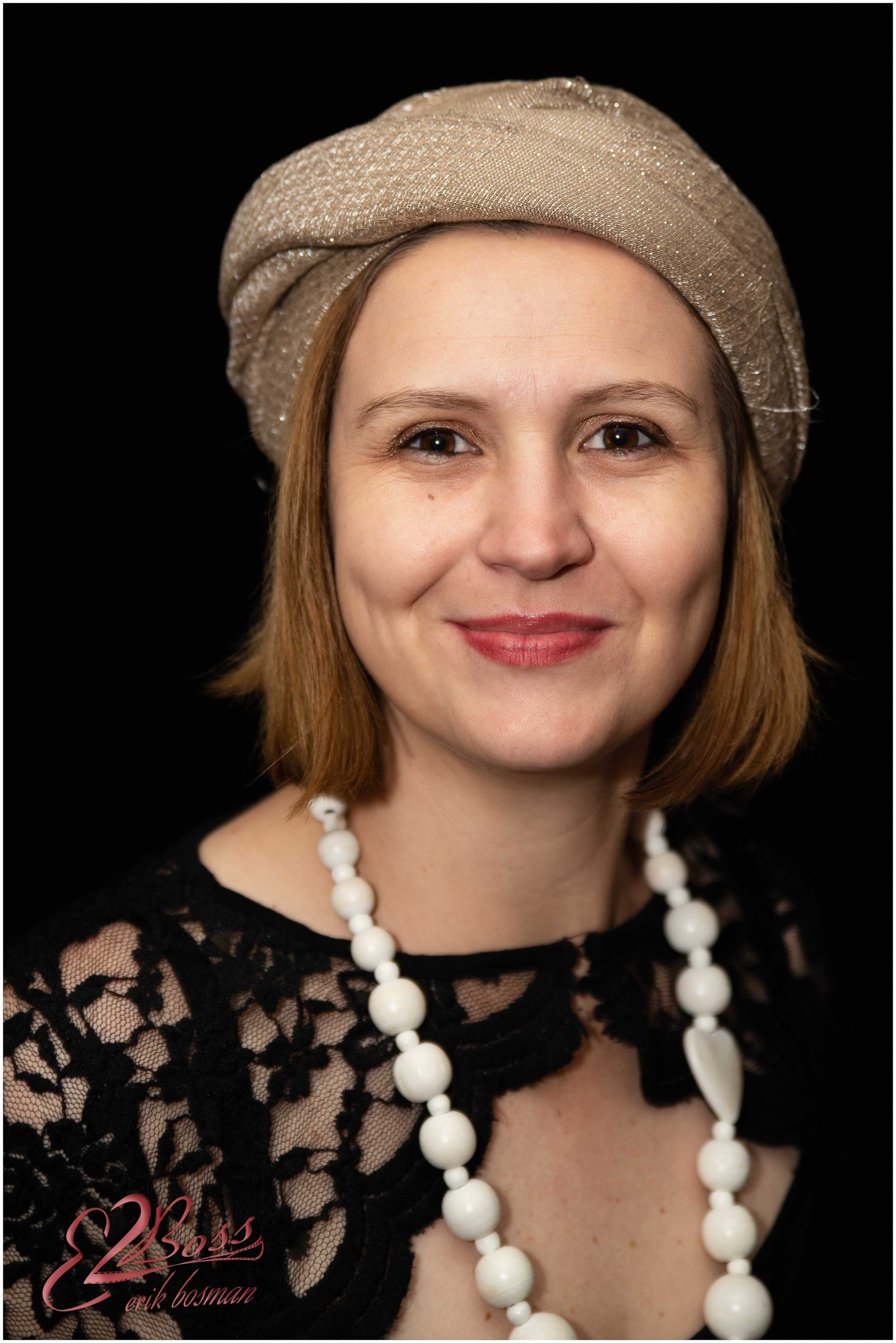 A woman with short brown hair wearing a beige head wrap, black lace top, white beaded necklace, and red lipstick, smiling against a black background.