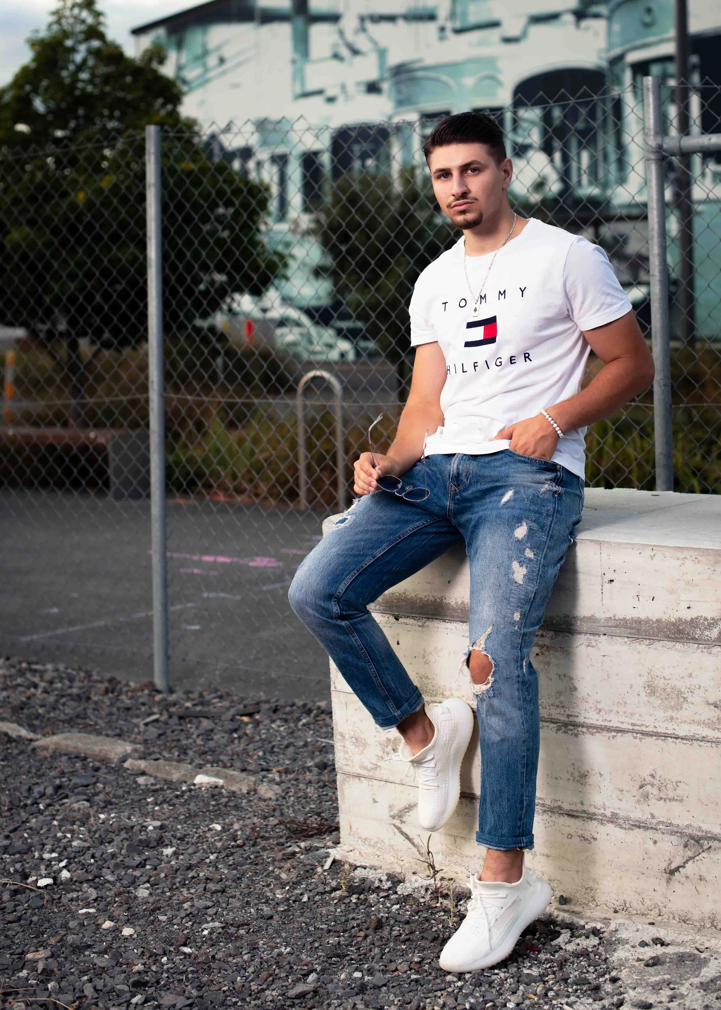 A young man with dark hair and a beard sitting on a concrete ledge in an outdoor urban setting, holding sunglasses, wearing a white Tommy Hilfiger t-shirt, distressed jeans, and white sneakers, with a chain bracelet on his wrist.