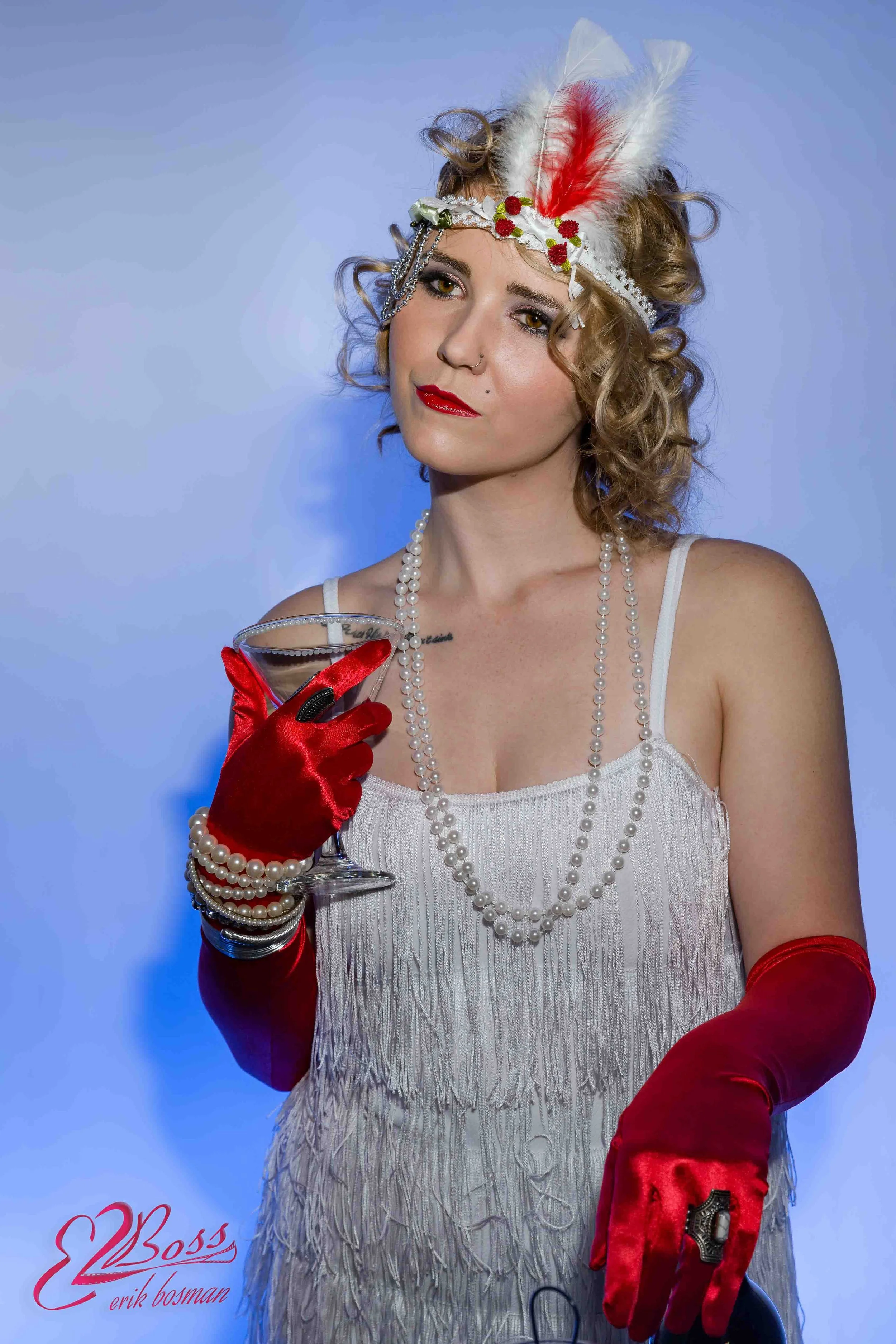 A woman with curly blonde hair wearing a feathered headband, multiple pearl necklaces, and red gloves, holding a cocktail glass and posing in front of a blue background.
