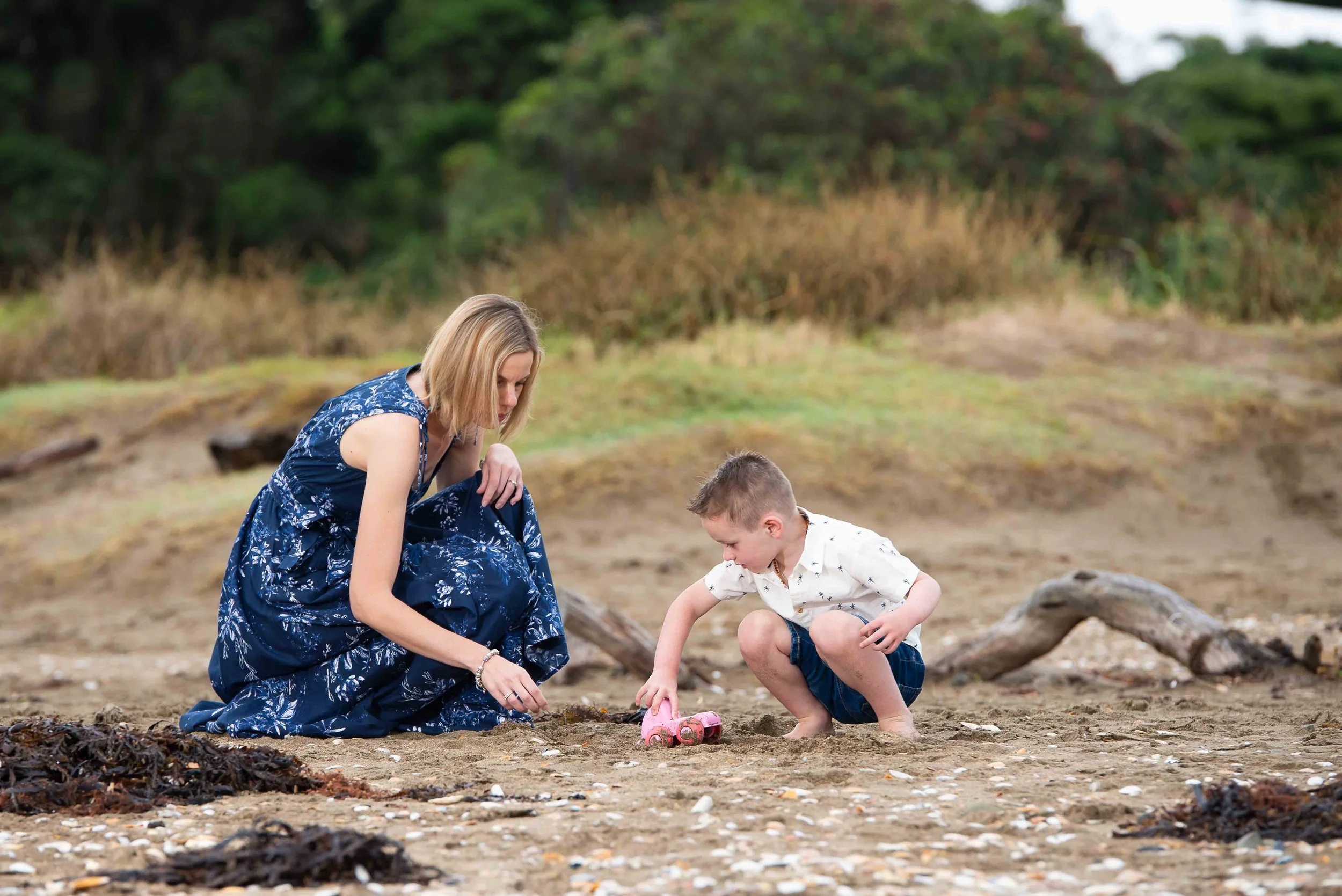 A woman and a young boy at the beach playing in the sand with a small pink toy car, surrounded by driftwood and seaweed.
