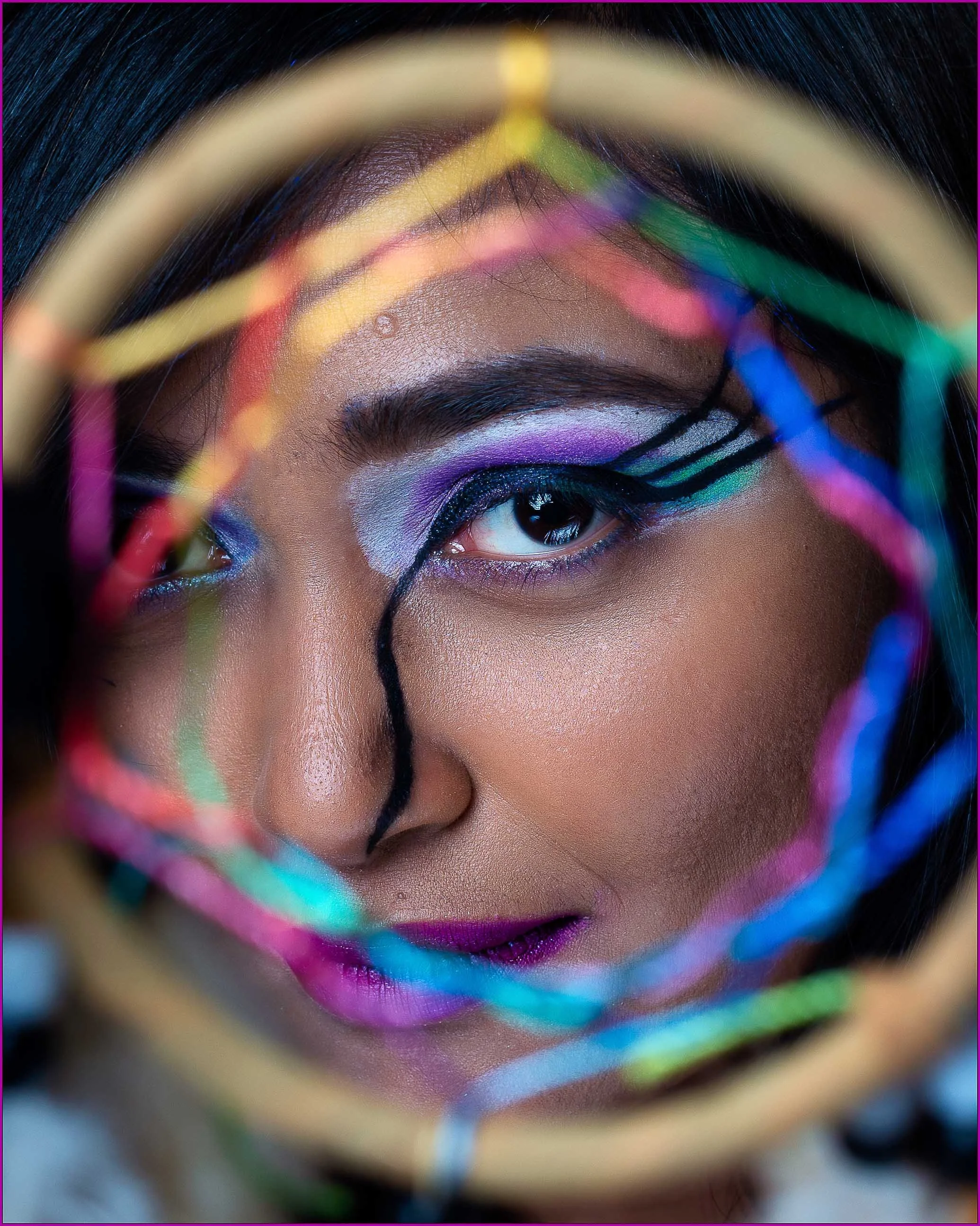 Close-up of a person's face with vibrant, artistic makeup, framed through colorful, intertwined strings.