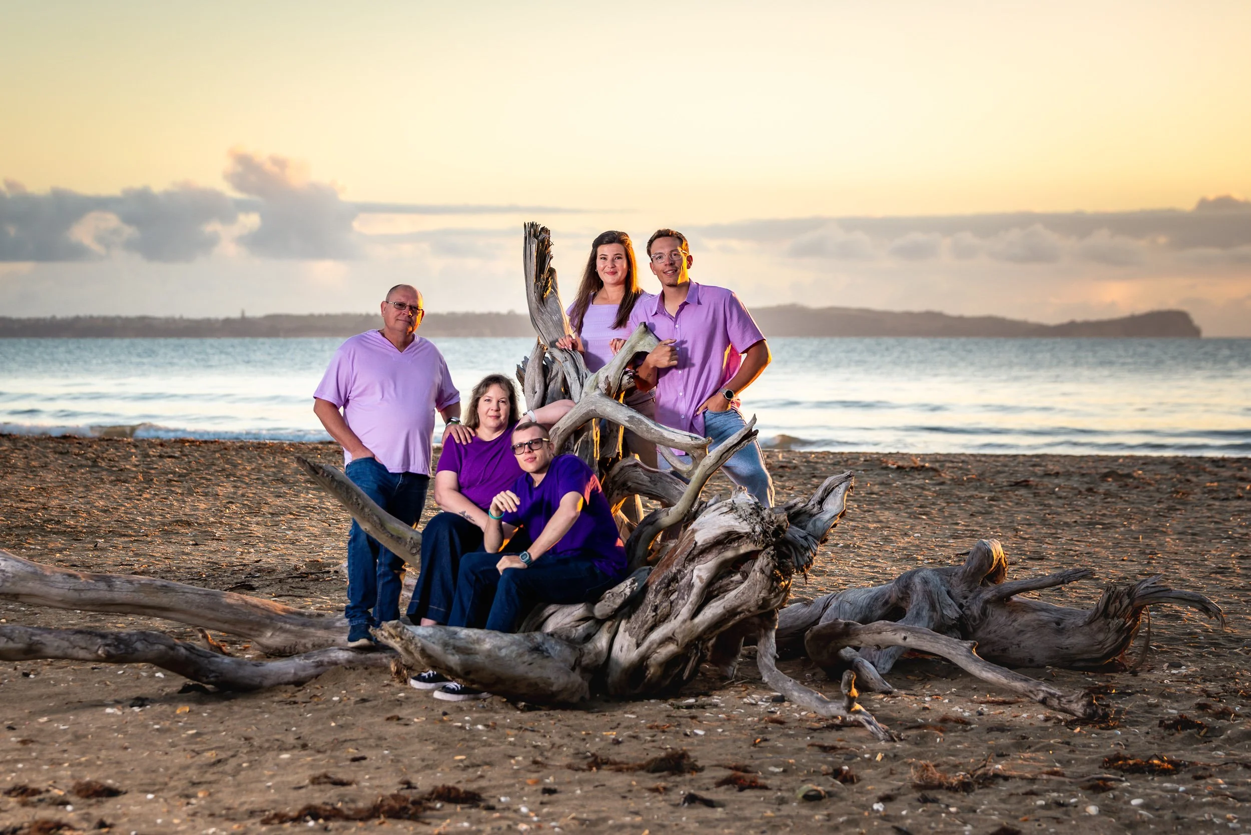 Family of five on the beach, with a driftwood sculpture and an ocean background during sunset, three standing and two sitting.