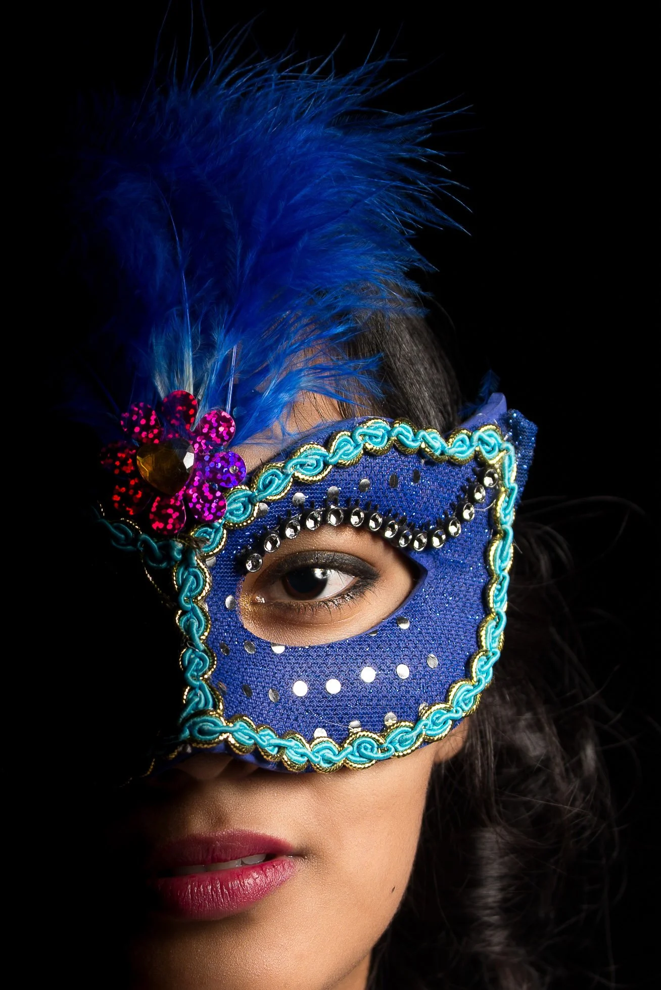 Close-up of a woman wearing an ornate blue masquerade mask decorated with beads, sequins, and embroidery, adorned with a large blue feather and a pink flower with a gem, against a black background.
