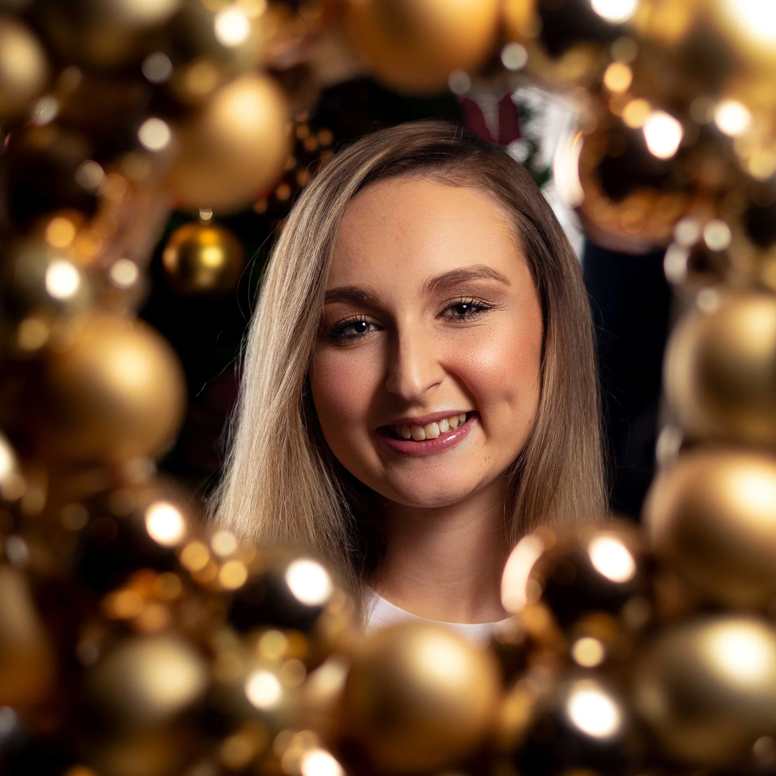 A young woman with blonde hair smiling through a circular opening decorated with gold Christmas ornaments.
