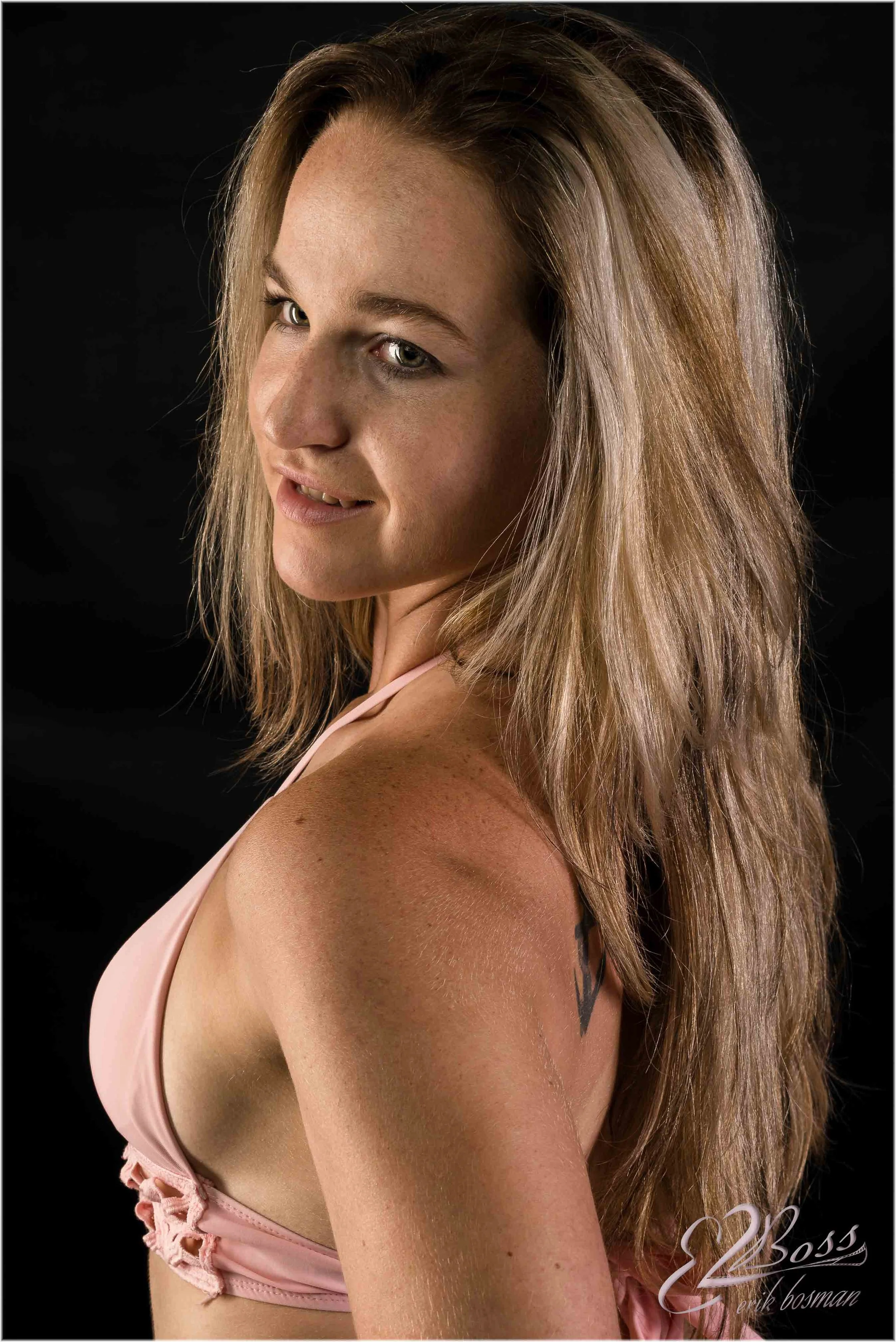 Portrait of a woman with long light brown hair and light skin, wearing a pink top, against a black background.