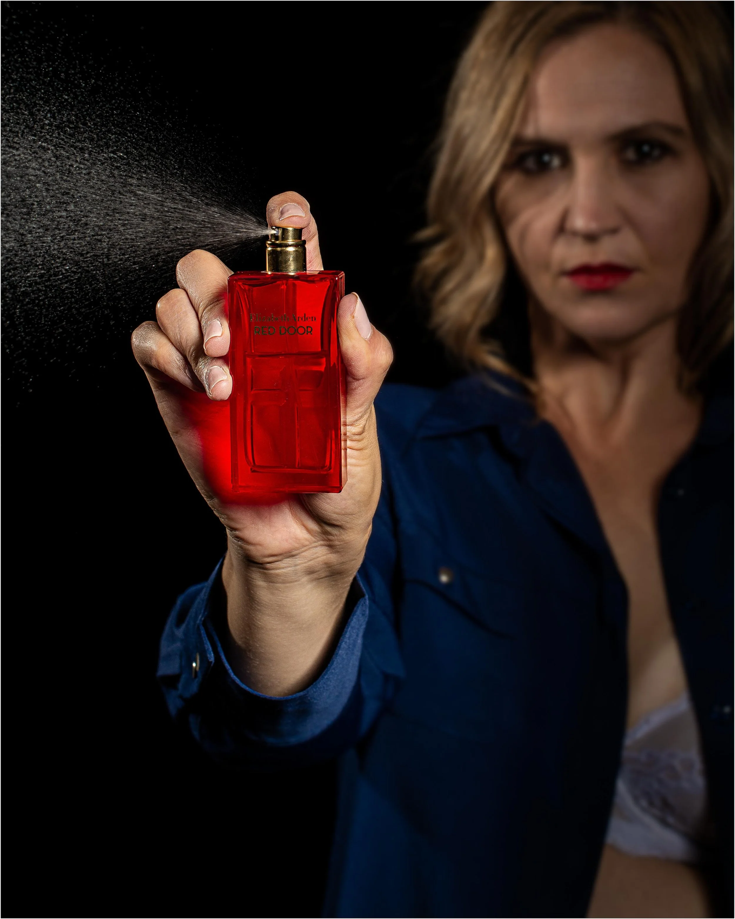 A woman with blonde hair and red lipstick holding a red perfume bottle labeled 'Elizabeth Arden Red Door,' spritzing perfume against a black background.