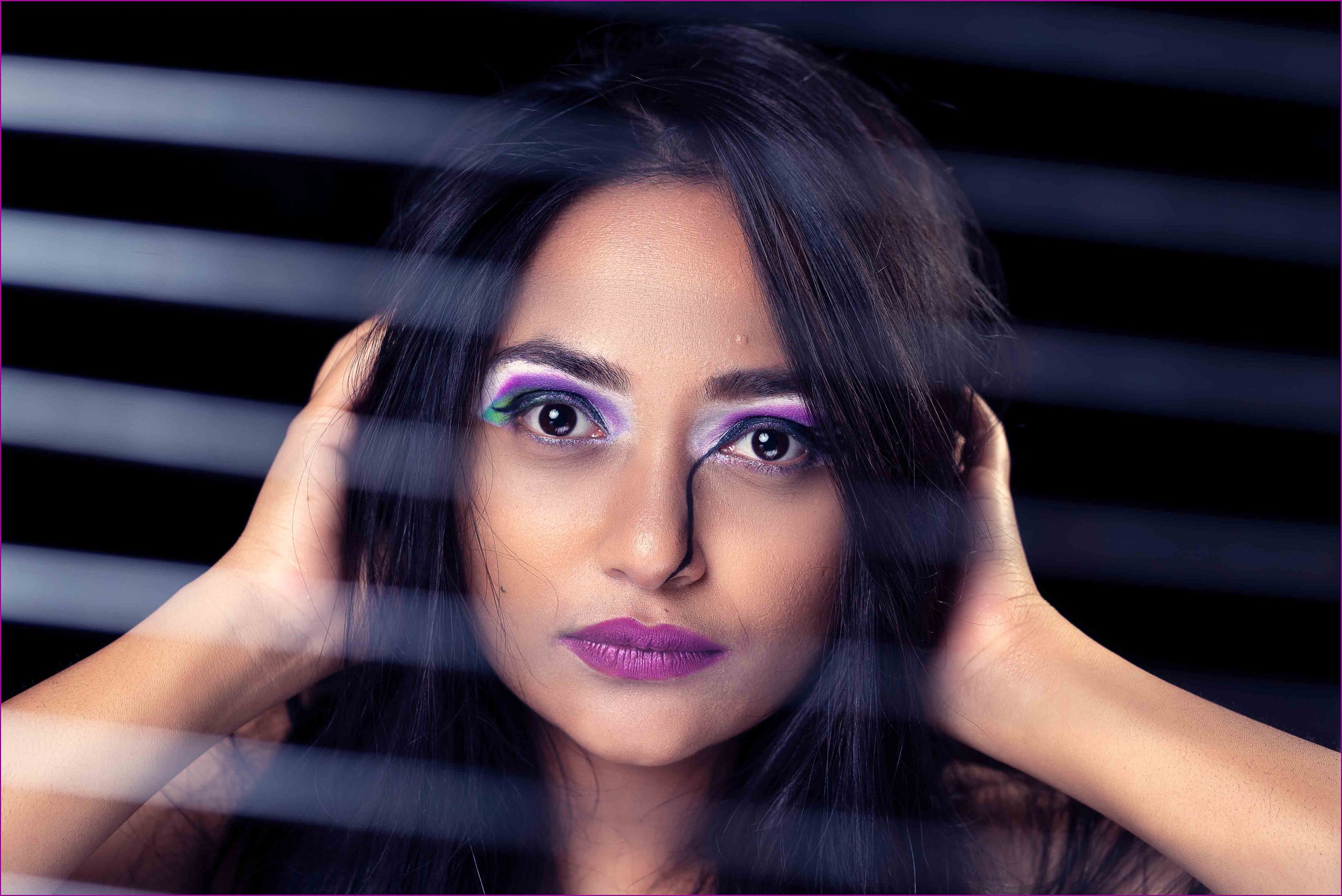 A young woman with long dark hair and vibrant colorful eye makeup looking through window blinds.