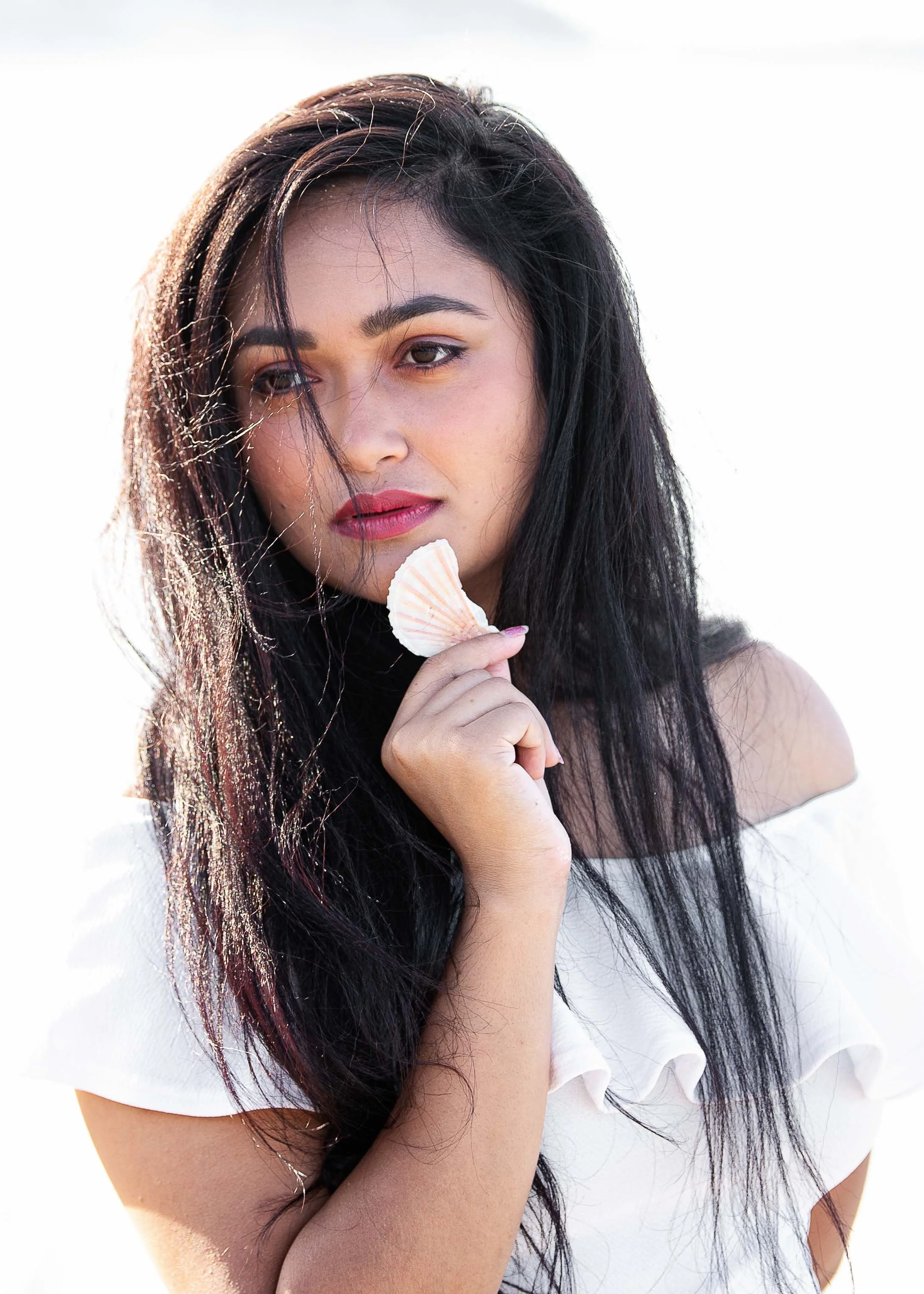 A young woman with long black hair holding a seashell near her chin, wearing an off-the-shoulder white top, with a neutral expression.