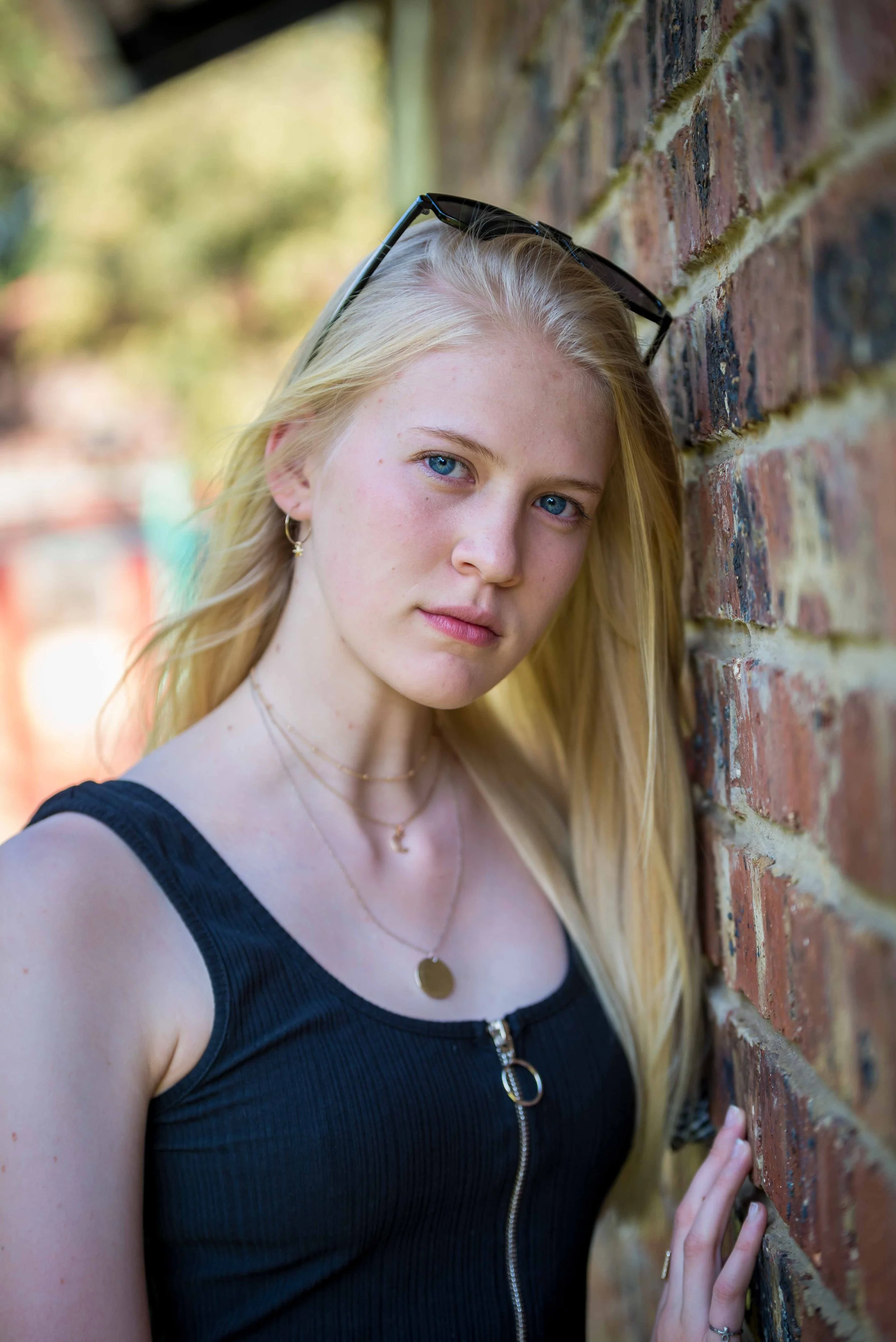 A young woman with blonde hair and blue eyes leaning against a brick wall outdoors, wearing sunglasses on her head, a black zippered top, and gold jewelry, with a blurred green background.