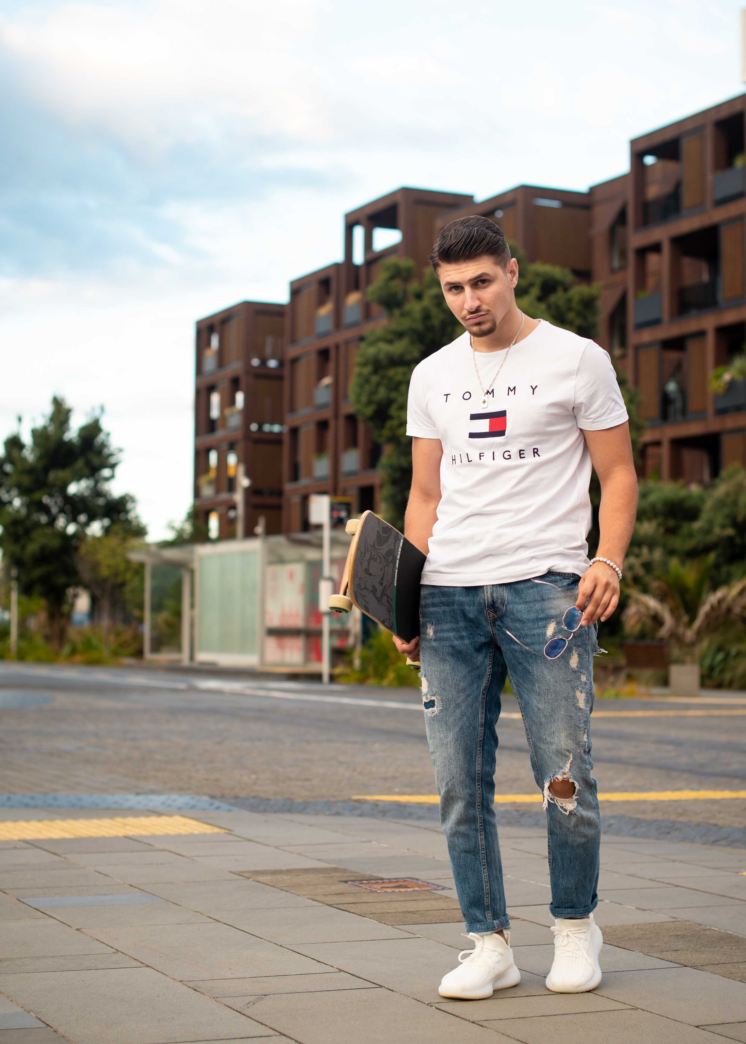 Young man in casual clothing holding skateboard standing on sidewalk in urban area