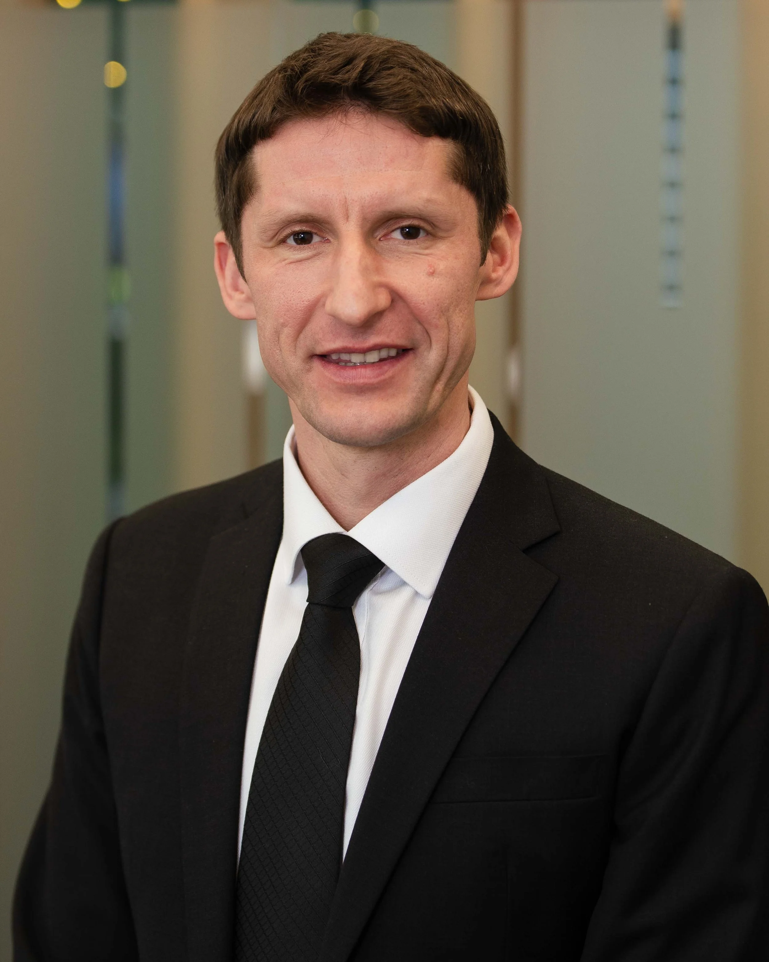 A man in a black suit, white shirt, and black tie standing in front of a blurred background with lockers.