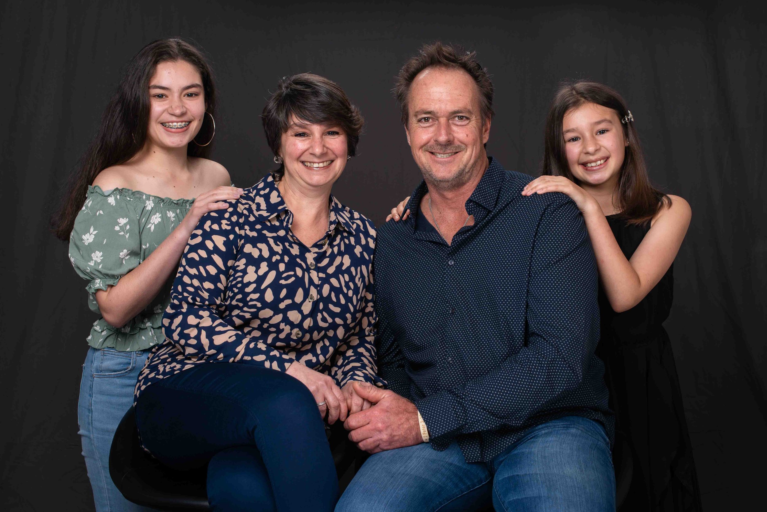 A family of four posing together against a dark background. The mother and father are seated in the front, holding hands, while the two daughters stand behind them with their hands on the parents' shoulders. All are smiling.