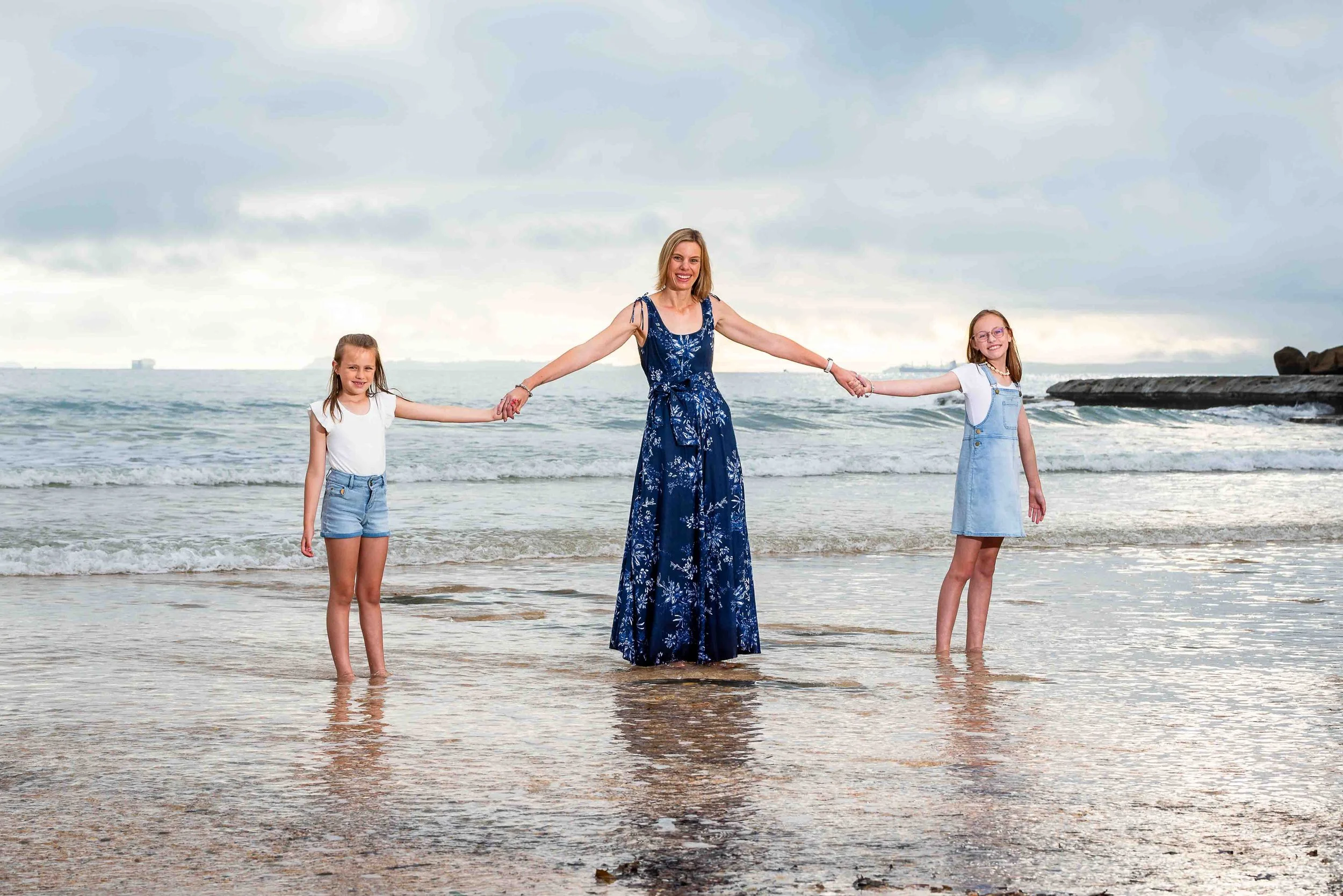 A woman and two young girls holding hands on a beach with ocean waves and cloudy sky in the background.