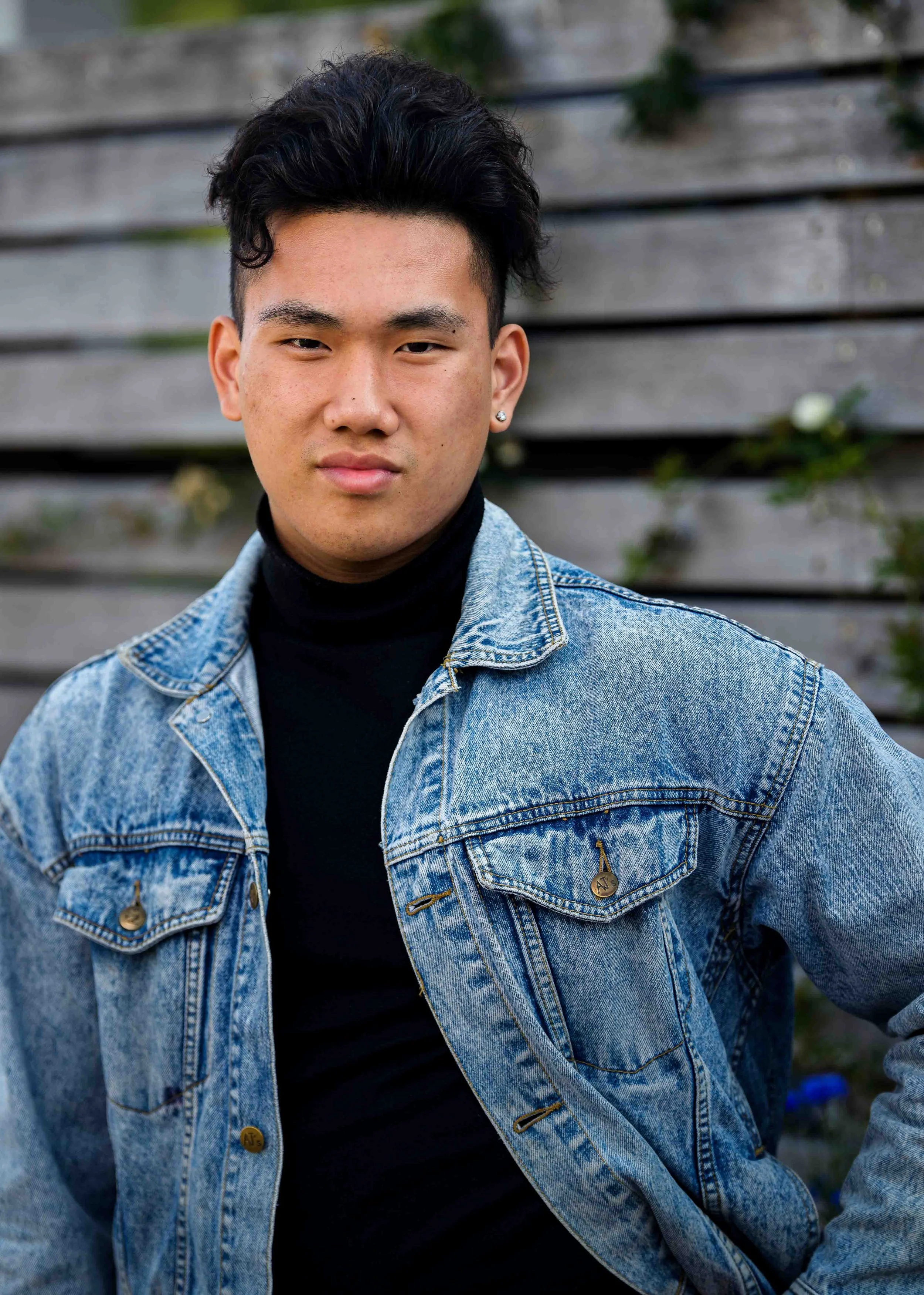 A young man with dark, short, wavy hair and earrings, wearing a black turtleneck and a light blue denim jacket, standing outdoors in front of a wooden fence.