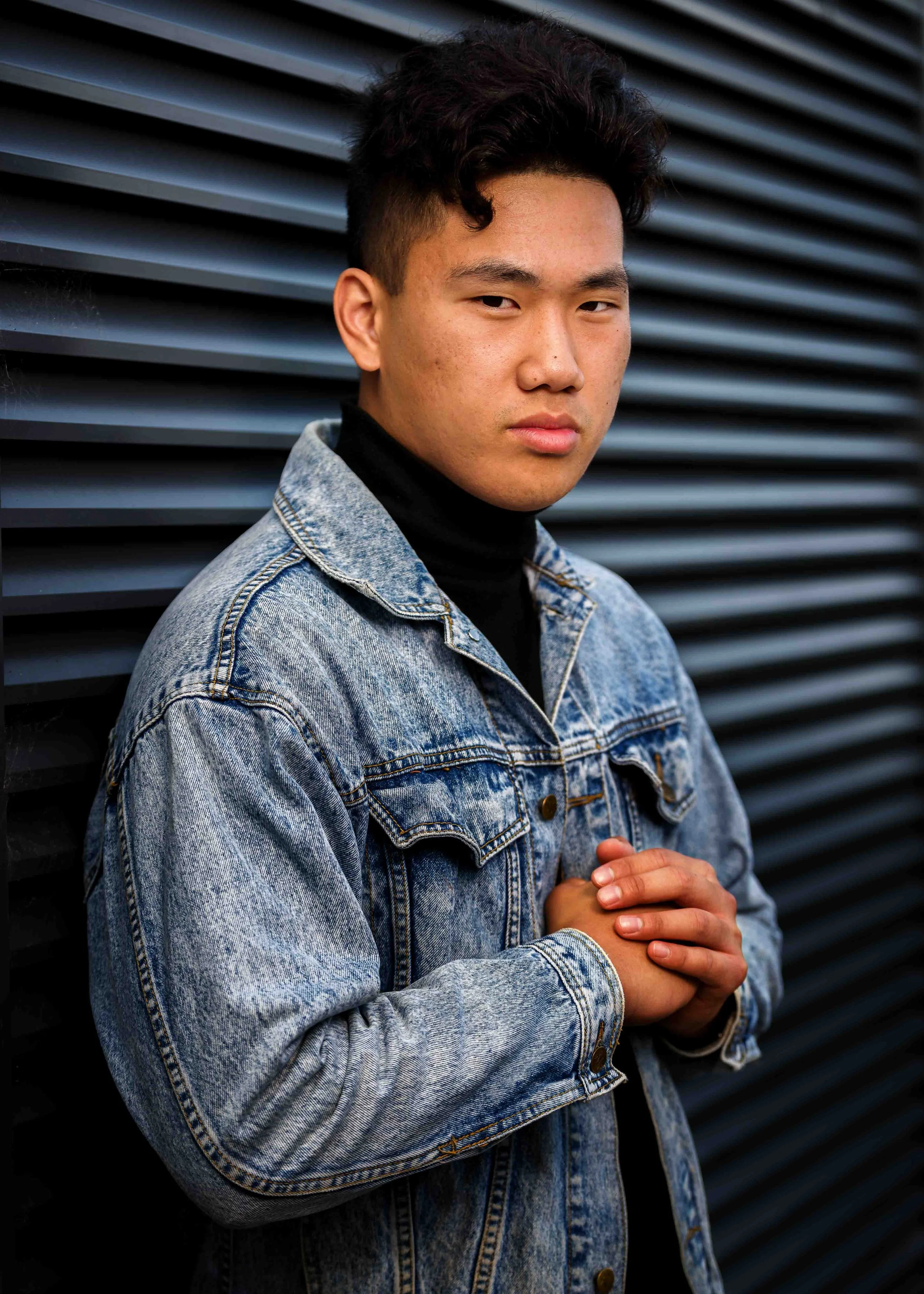 A young man with dark curly hair and wearing a denim jacket and black turtleneck stands against a dark, diagonal striped background.
