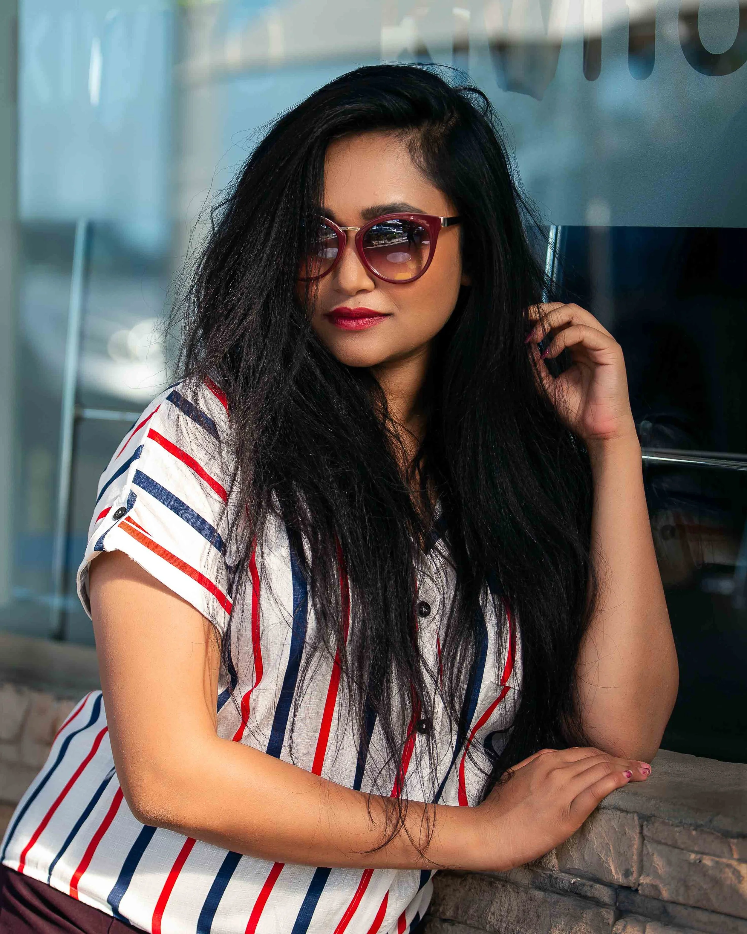 A woman with long black hair wearing sunglasses and a striped shirt posing outdoors.