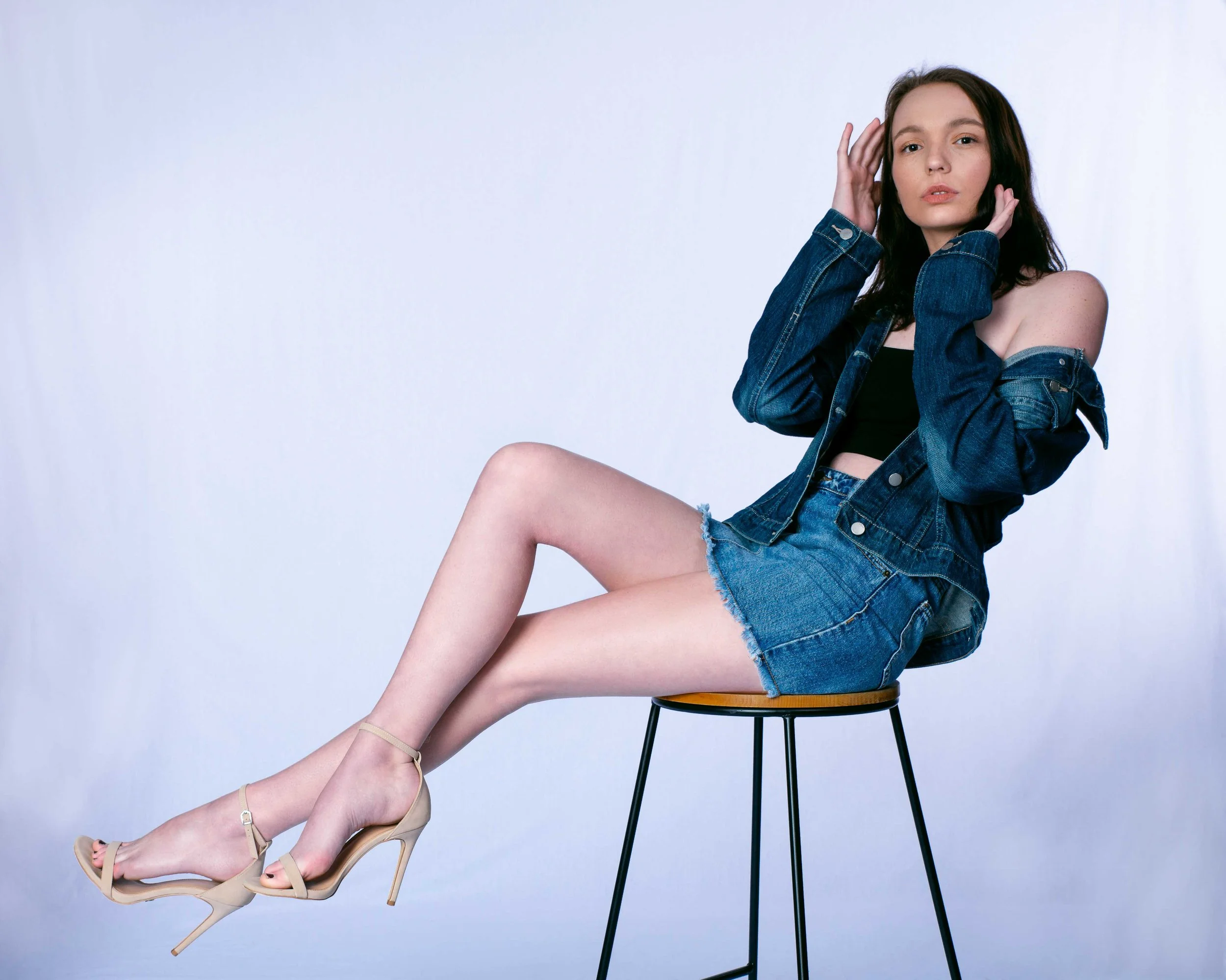 A young woman with long dark hair sitting sideways on a wooden stool, wearing a denim jacket off the shoulder, black crop top, denim shorts, and beige high-heeled sandals, with a neutral expression against a plain white background.