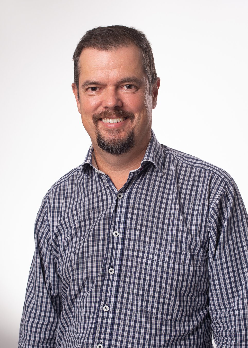 Portrait of a smiling man with short brown hair, beard, and mustache, wearing a checkered shirt, against a white background.