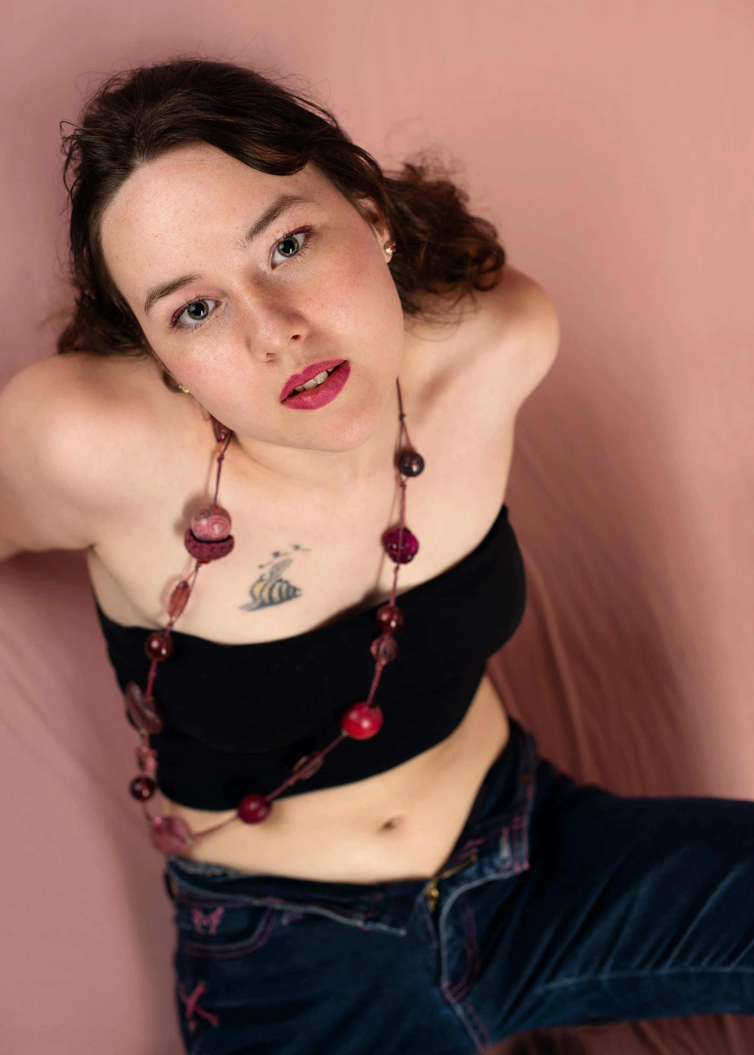 A young woman with dark hair and fair skin, wearing a black strapless top and dark jeans, is sitting against a pink background, looking up at the camera with a slightly open mouth. She has a necklace with large pink and red beads and a small tattoo o