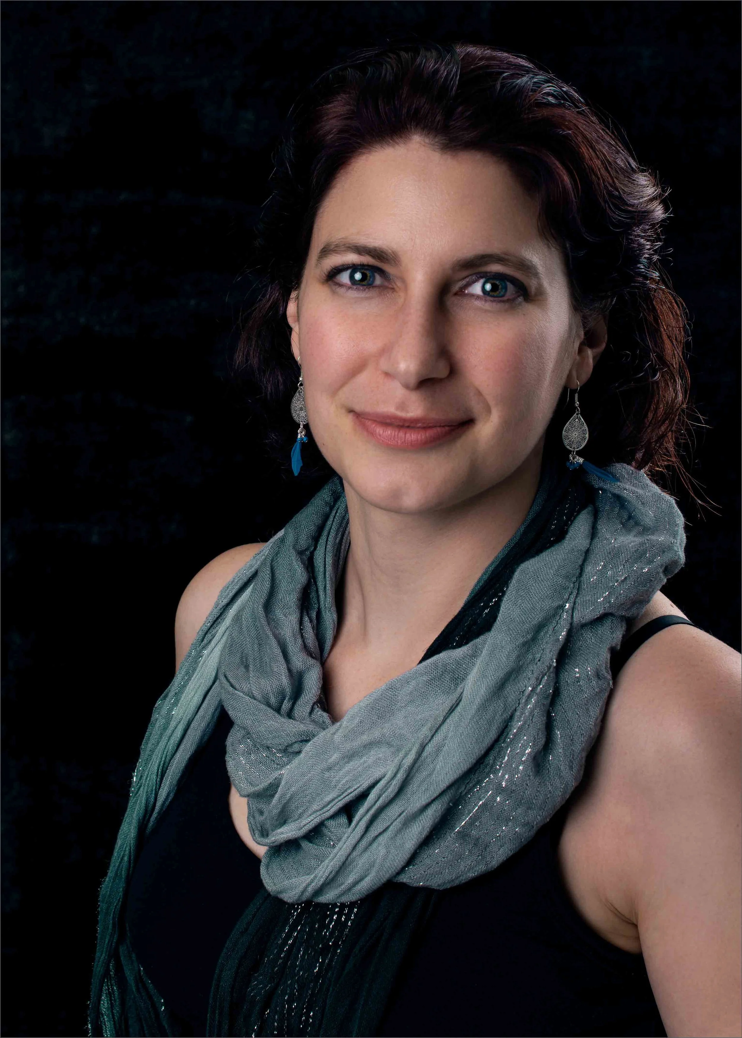 A woman with blue eyes, short dark hair, wearing earrings and a scarf, smiling at the camera against a black background.