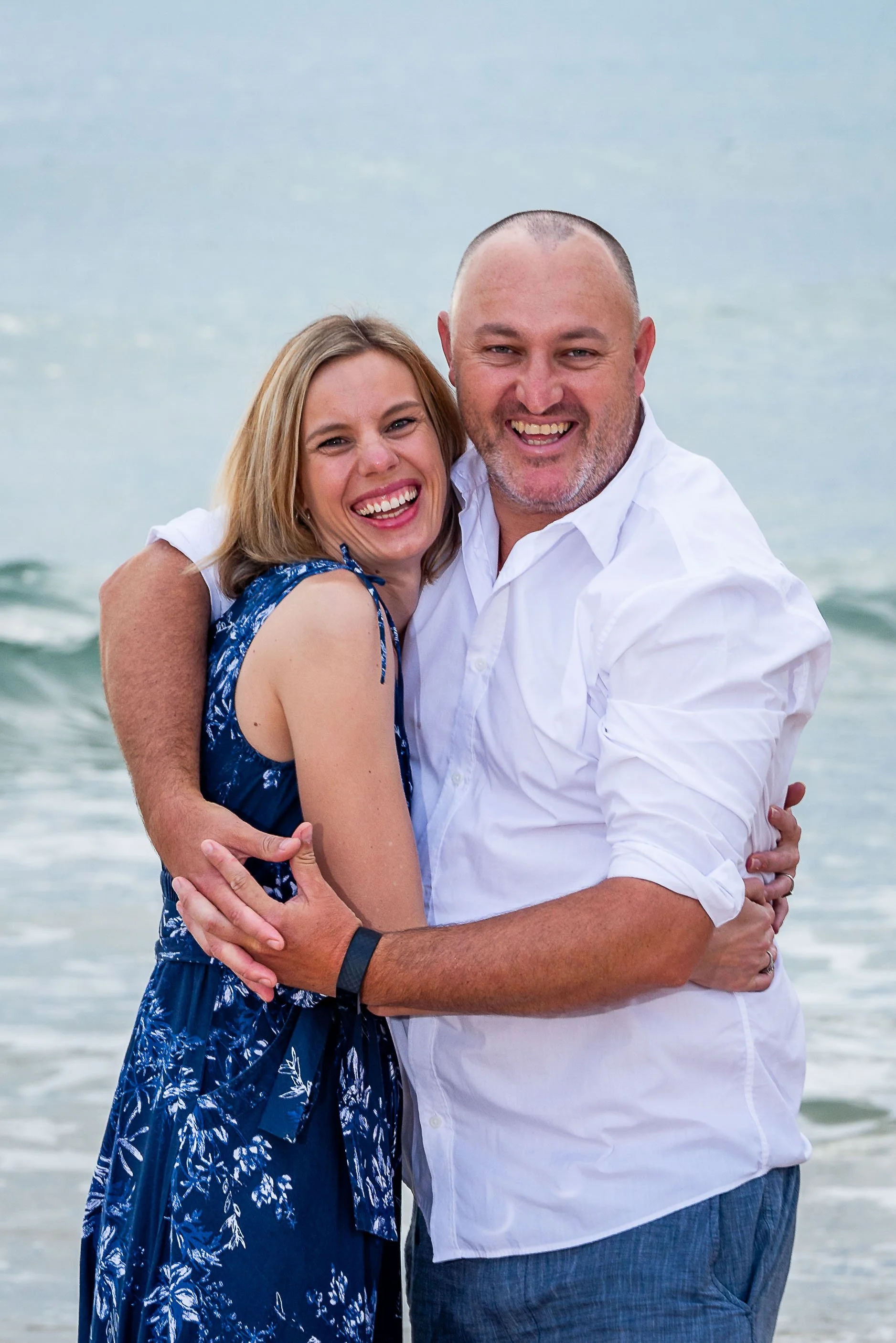 A smiling couple embraces on the beach with water and waves in the background, both wearing casual summer clothes.