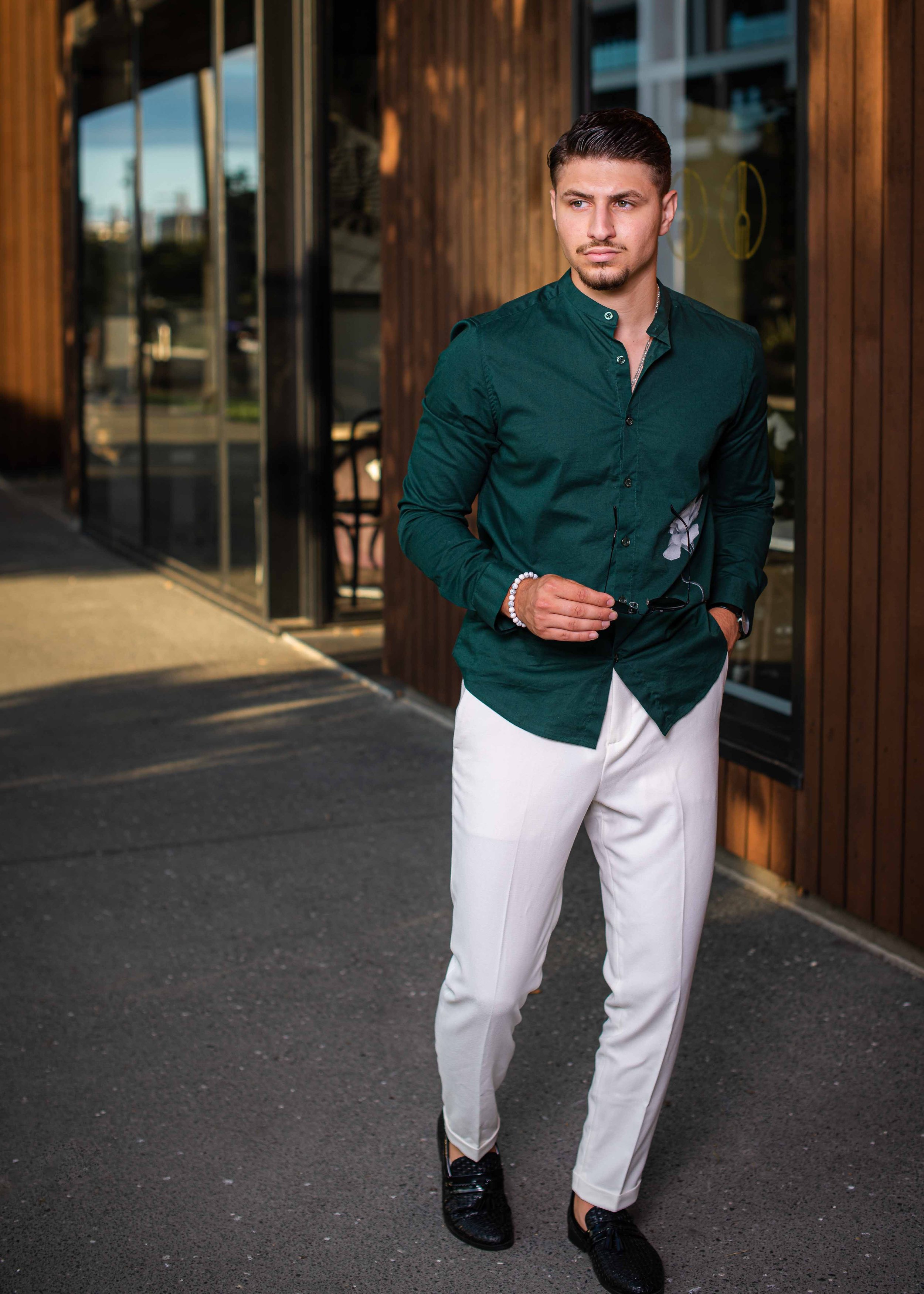 A young man in a green button-up shirt, white pants, black shoes, and sunglasses, standing outside a building with wooden exterior, during daytime.