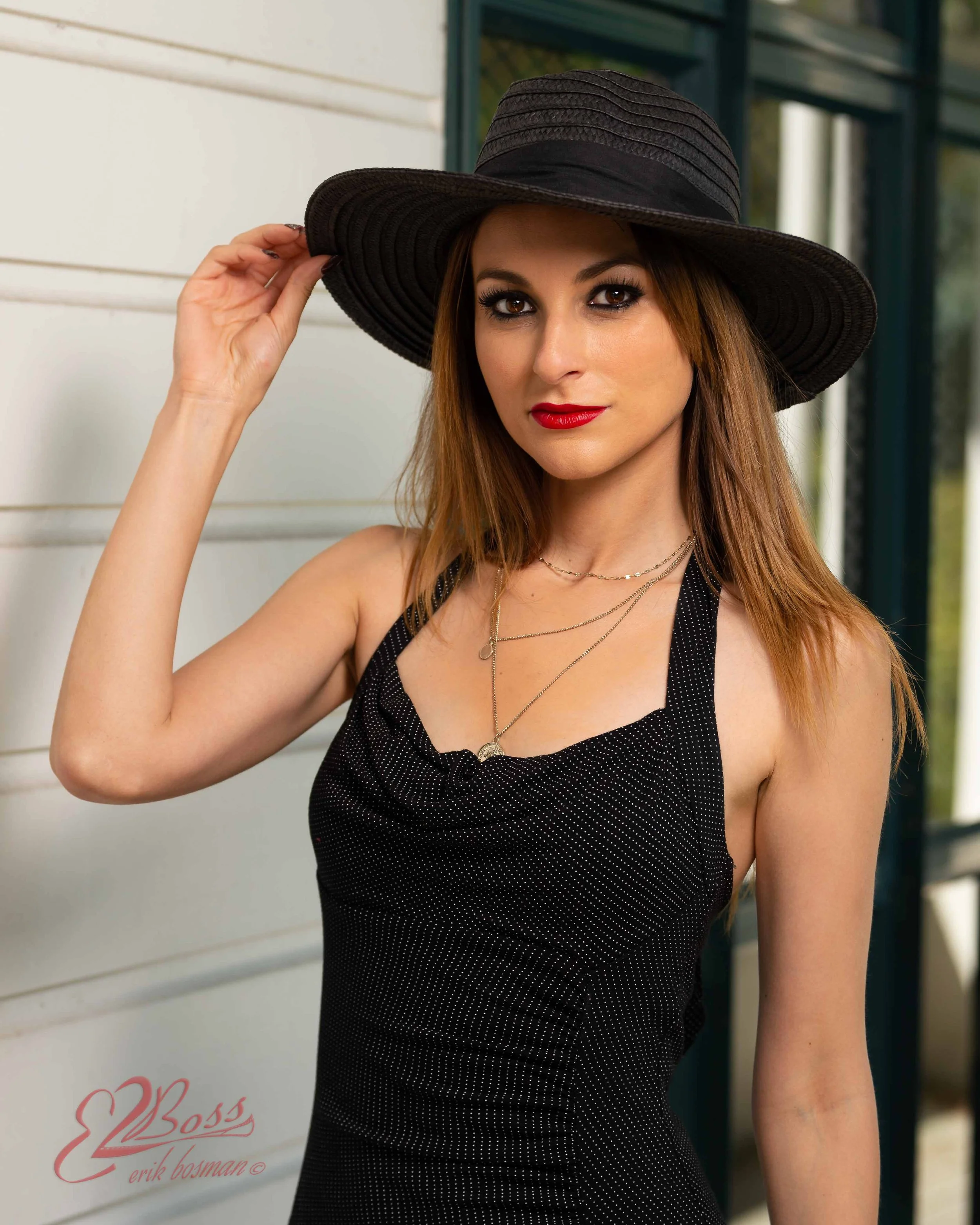 A woman with long, reddish hair wearing a wide-brimmed black hat, black sleeveless dress, and layered necklaces, posing outdoors next to a white wall and a green window frame.